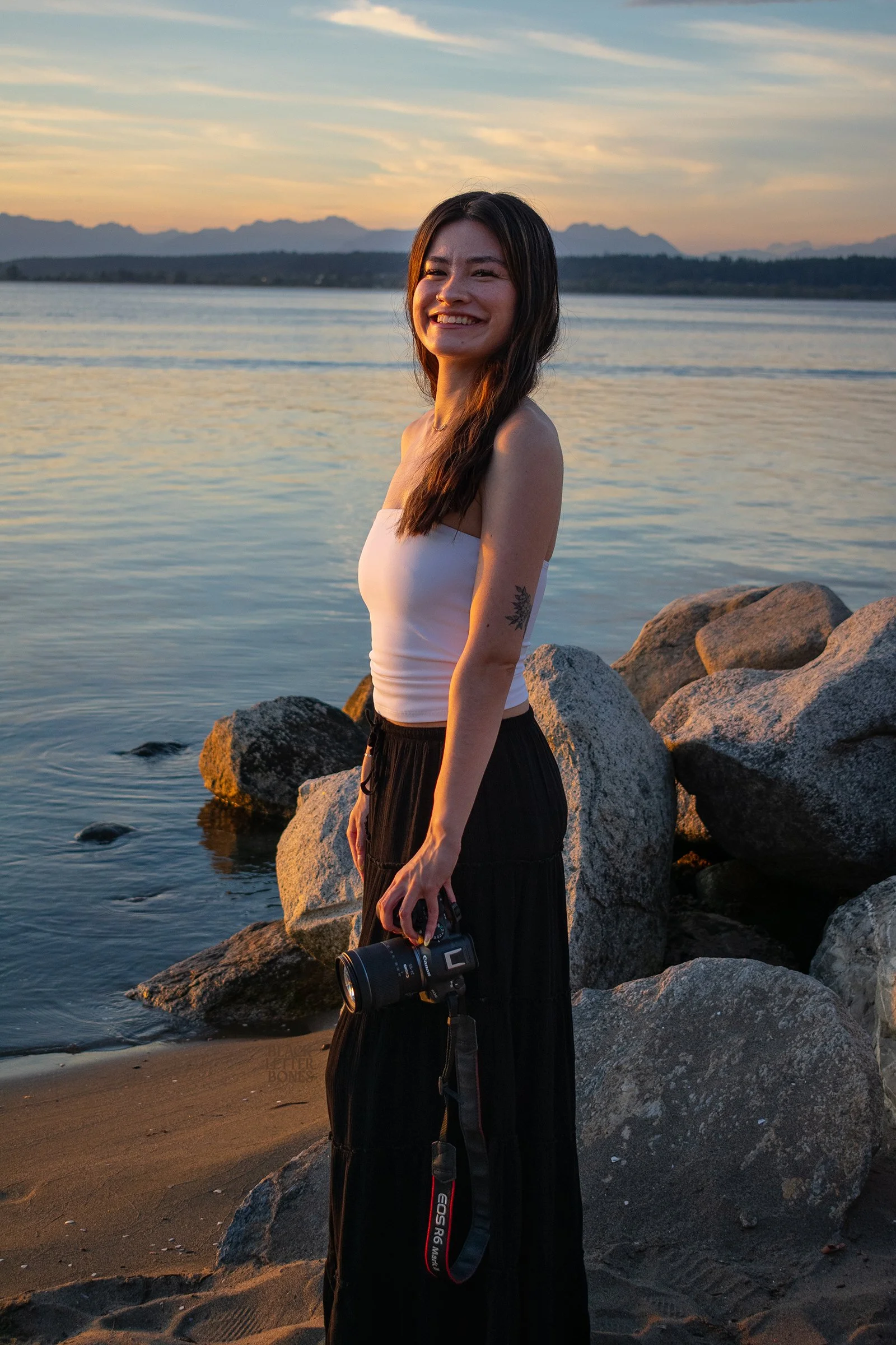 A woman standing on a beach near rocks during sunset, holding a camera, wearing a white tube top and black pants, smiling.