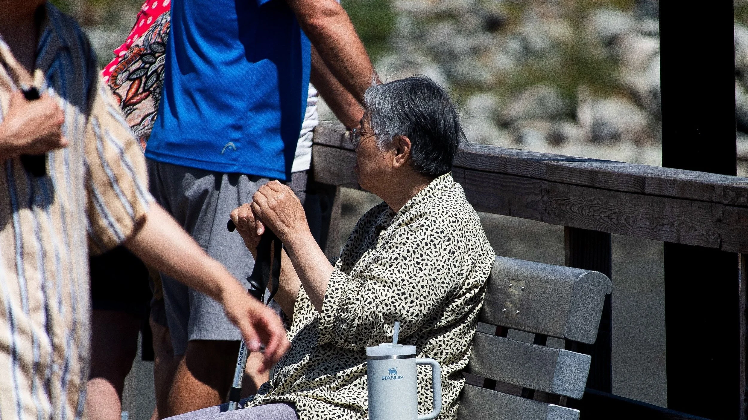 An elderly person with gray hair and glasses, wearing a animal print shirt, sitting on a bench with a thermos and straw in front of them, surrounded by a group of people outdoors with a rocky background.