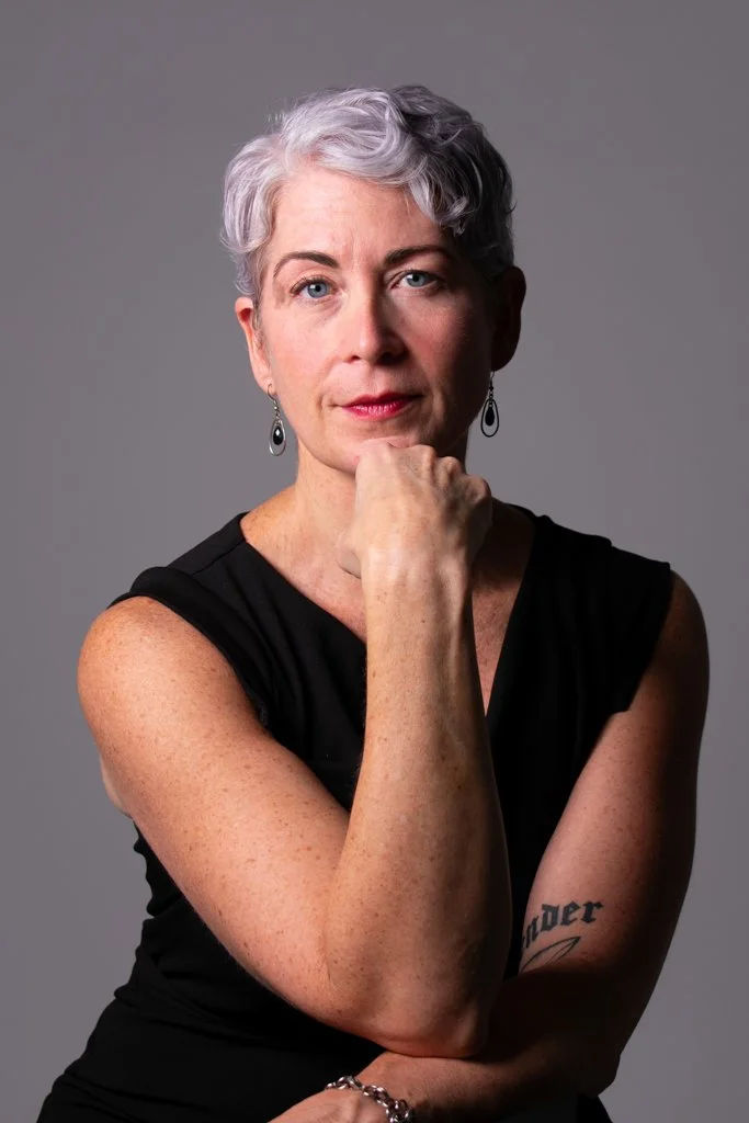 A woman with short gray hair, wearing a black sleeveless top, silver earrings, and a bracelet, poses with her hand on her chin against a plain gray background.