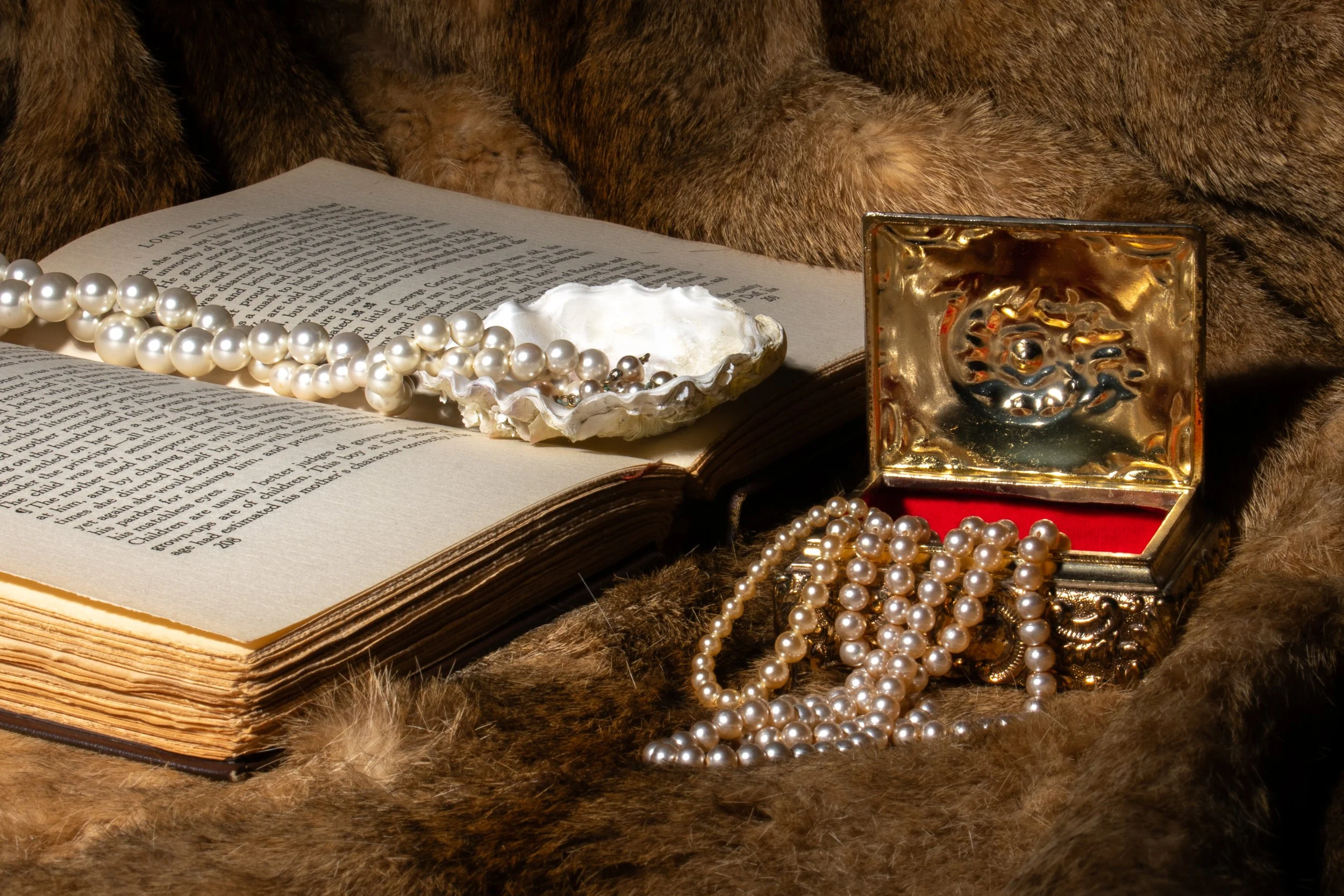 An open vintage Bible with pearl necklaces and a large oyster shell resting on its pages, surrounded by a jewelry box and a fur fabric background.