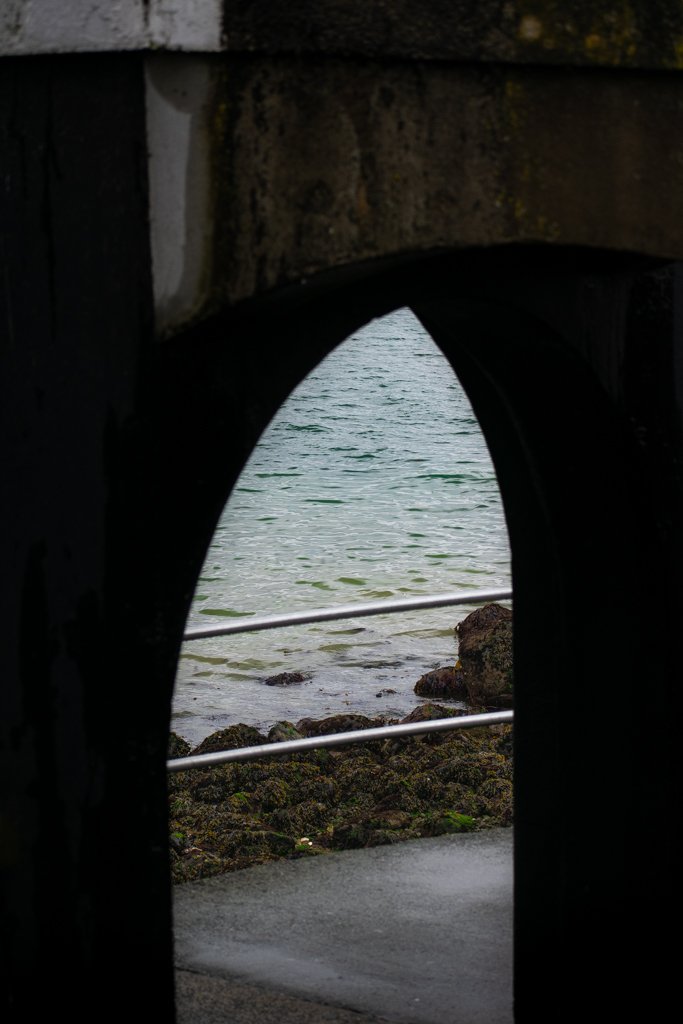 View of the ocean or sea through a small, arched opening in a stone or concrete structure, with rocks and water visible outside.
