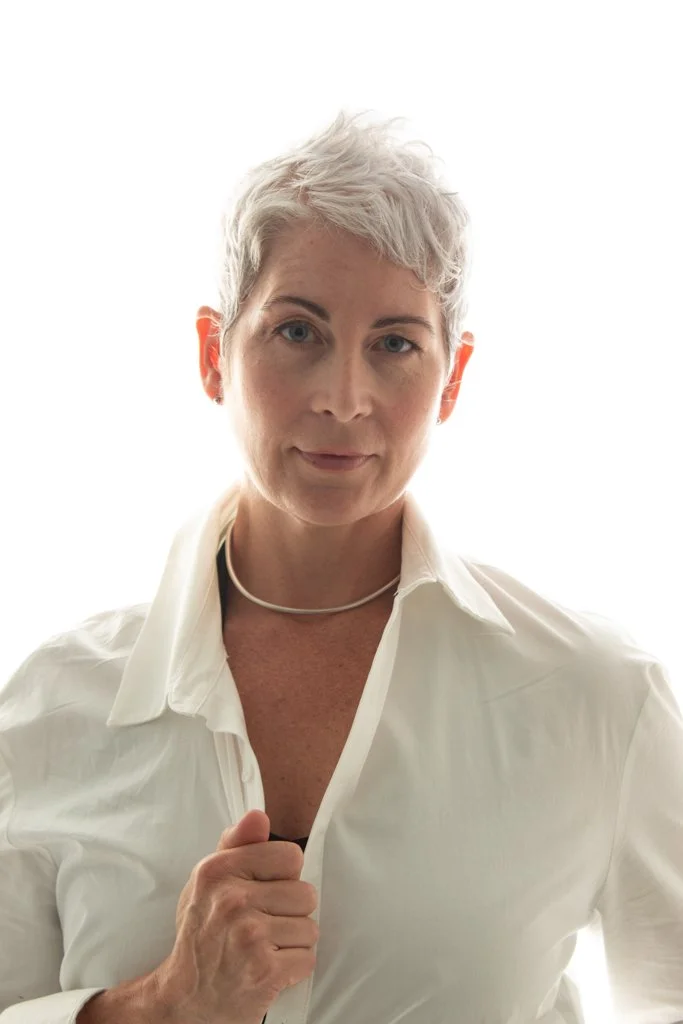 A woman with short, gray hair wearing a white button-up shirt and a silver necklace, looking directly at the camera with a slight smile.