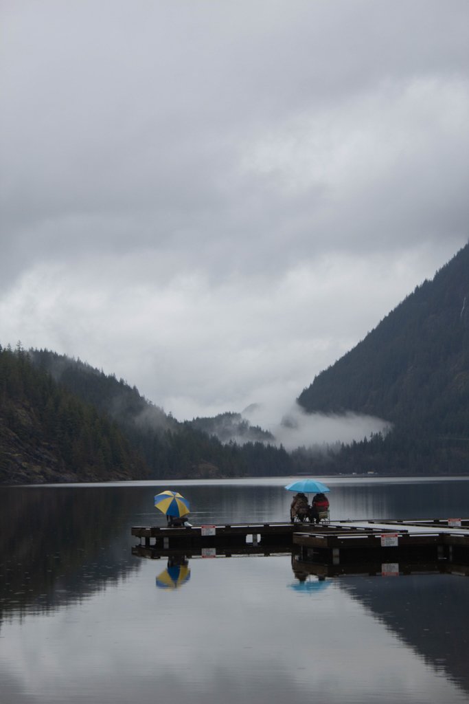 People sitting on a dock by a calm lake with umbrellas, surrounded by misty mountains and cloudy sky.