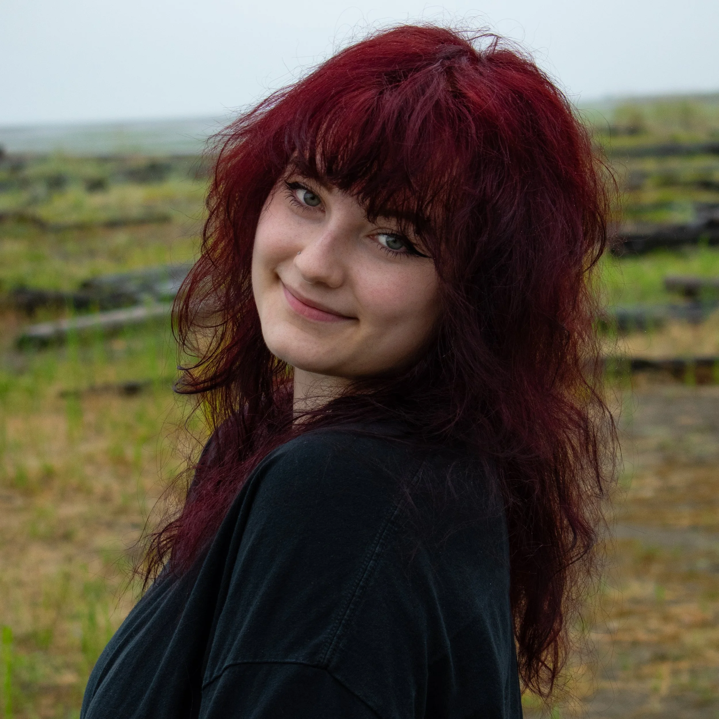 A woman with red, wavy hair and blue eyes smiling outdoors with a green, grassy background.