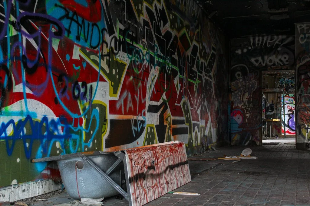 An abandoned building interior with graffiti-covered walls, a tipped-over trash bin, and debris on the floor.