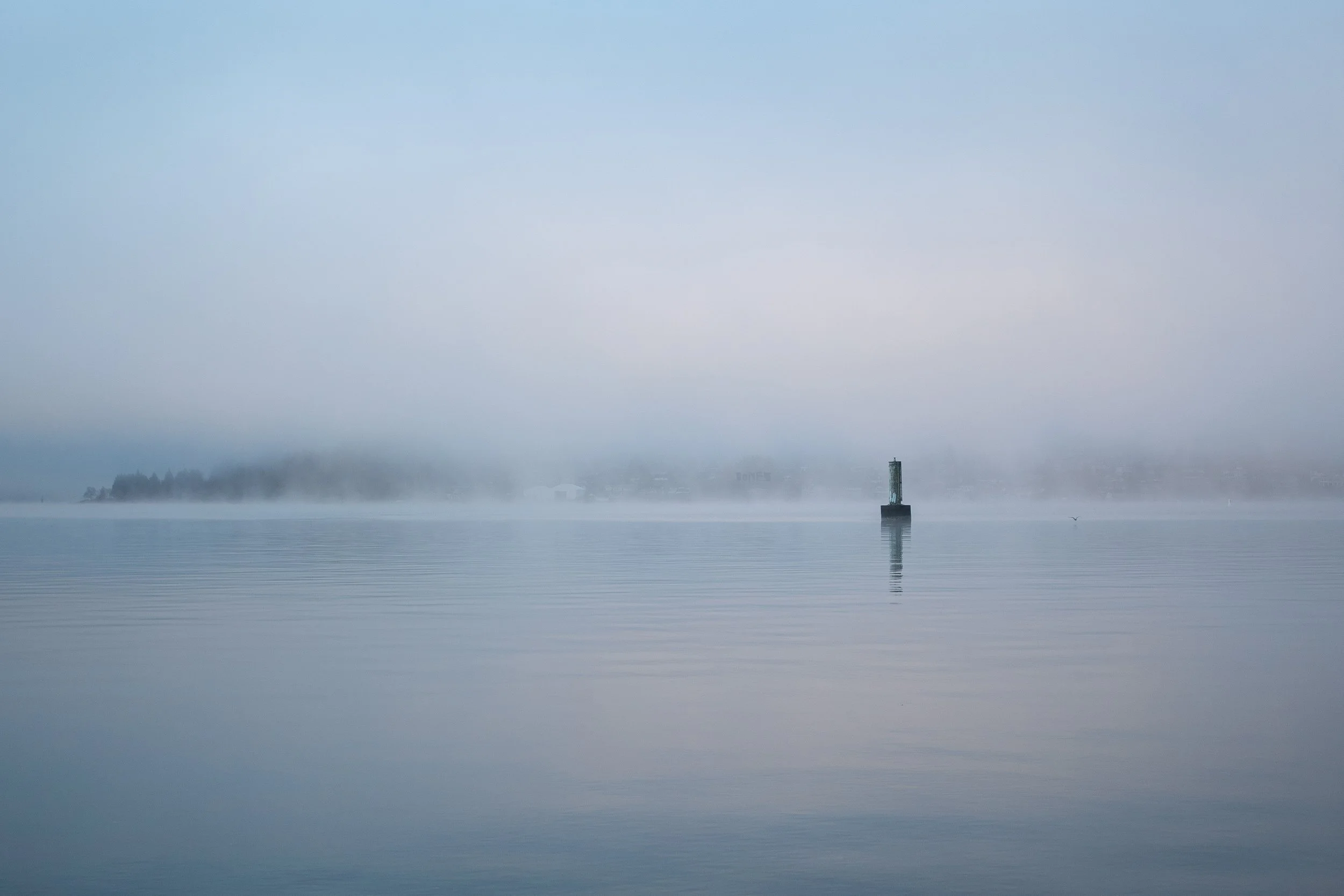 A foggy scene over a calm body of water with a small lighthouse on a pier in the distance and a faint shoreline with trees in the background.