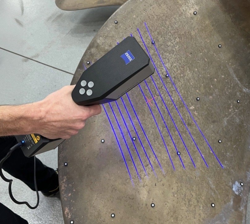 A person using a Zeiss 3D laser scanner to scan a metal surface with multiple small holes, revealing purple laser lines and some scattered metal beads on the surface.