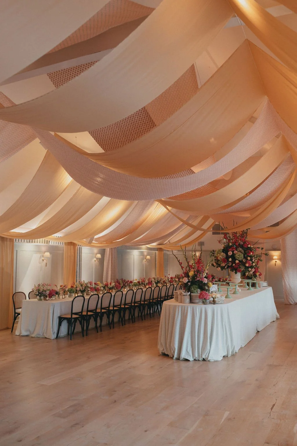 Elegant banquet hall decorated for a wedding or event with a long table adorned with floral arrangements, a separate dessert or gift table, and ceiling drapes in shades of beige and pink.