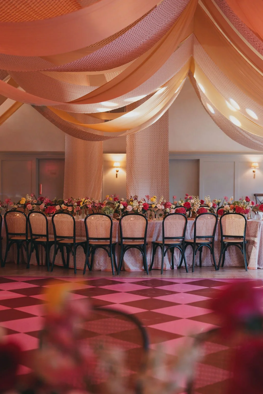 Elegant banquet hall with pink draped fabric ceiling, a long table decorated with pink, white, and red flowers, and surrounded by black chairs, in a softly lit room with checkered pink and gray flooring.