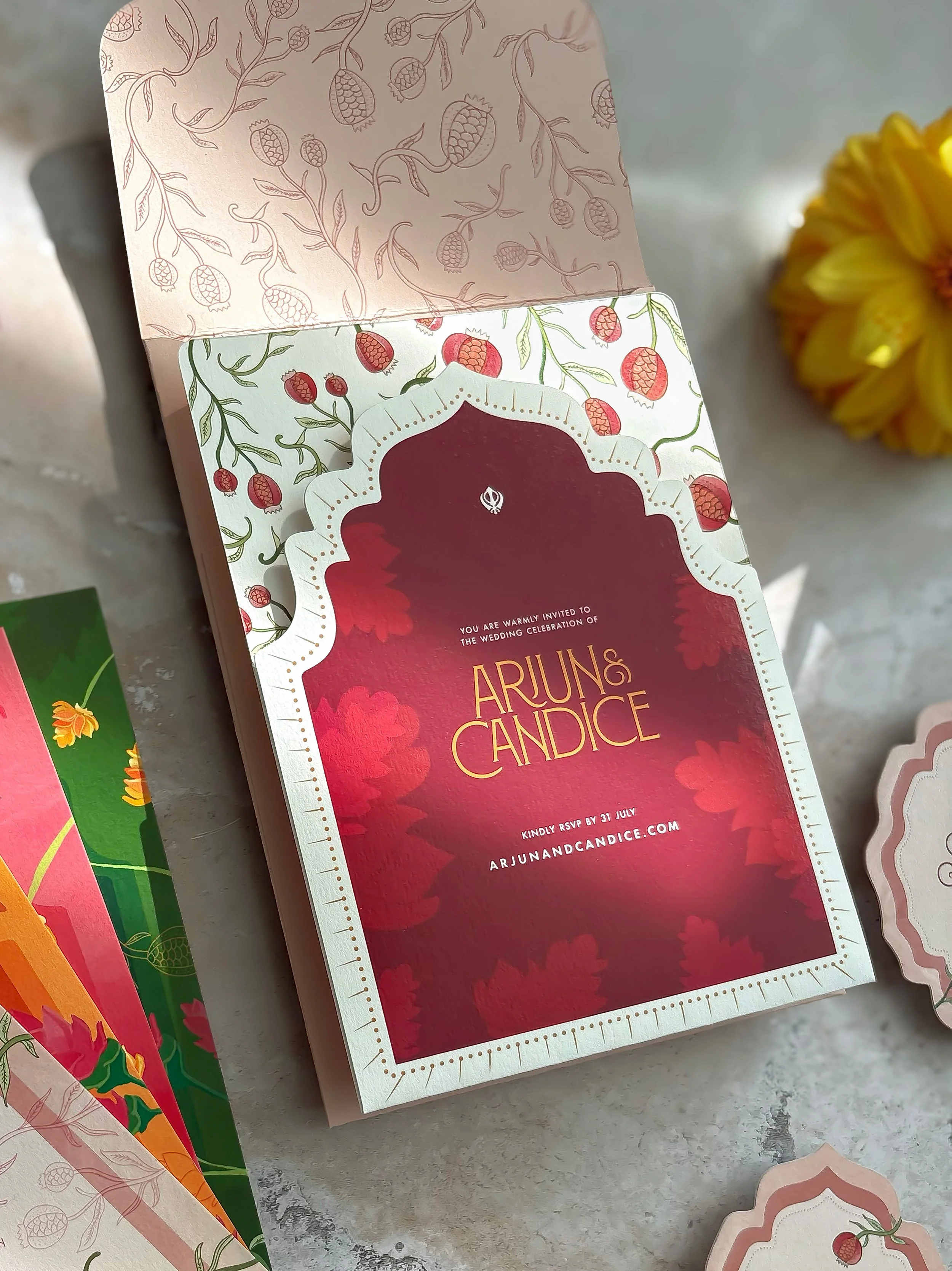 Invitation card for a wedding celebration of Arjun and Candice with a red background, white decorative border, and floral motif with hand-drawn pomegranates.
