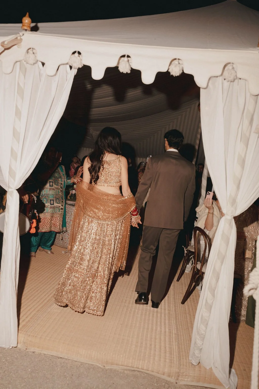 Couple walking hand in hand at a wedding reception, seen from behind through a white curtain, with guests in traditional and formal attire inside tent.