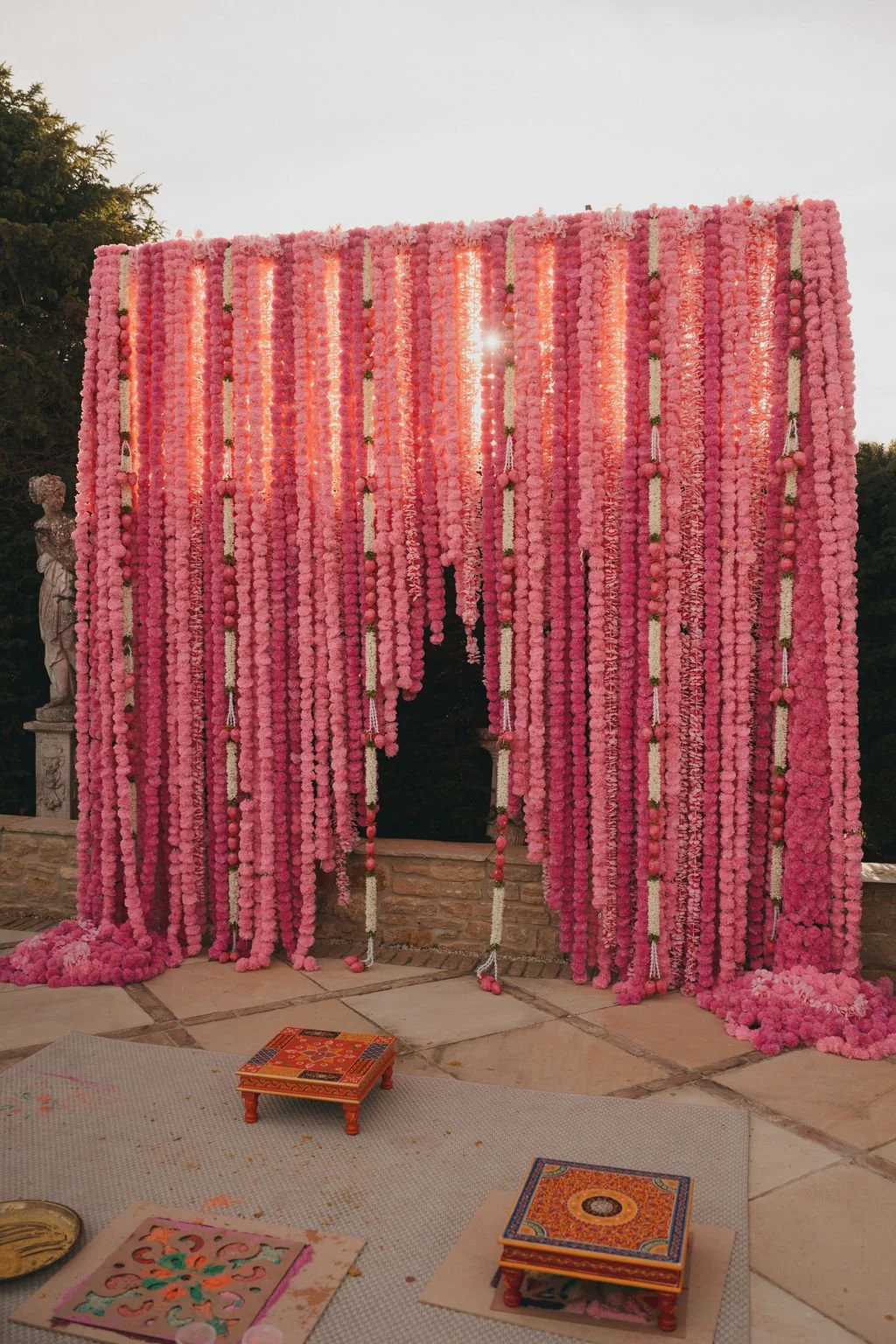 Pink floral wedding backdrop with hanging strings of flowers, illuminated by sunlight, with stone statues and decorated floor in the background.