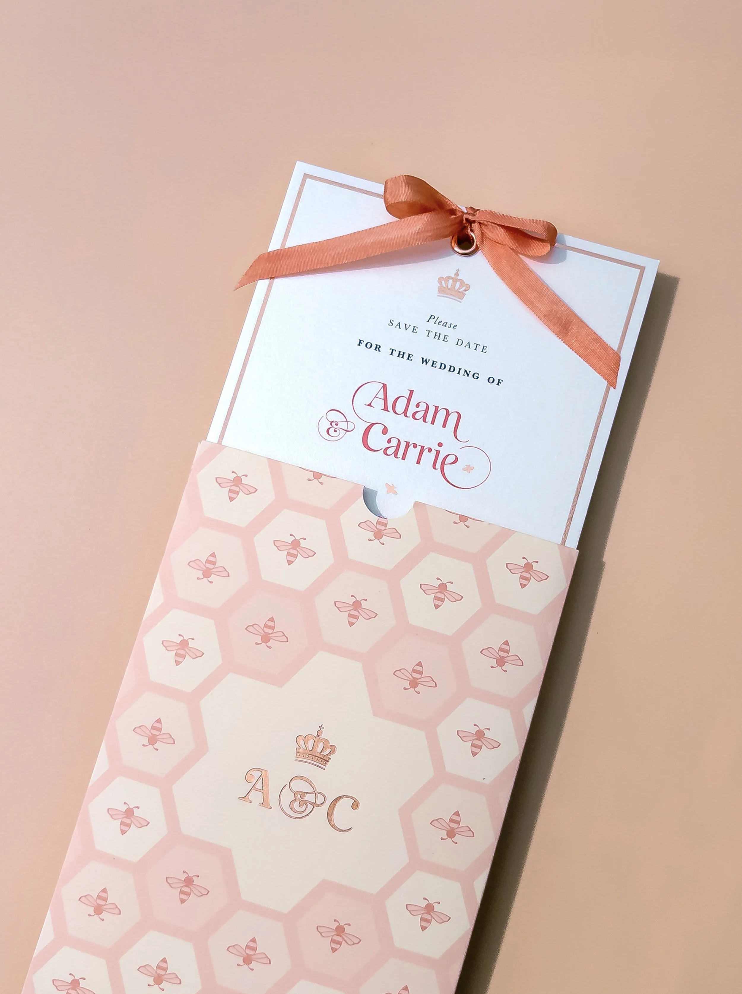 A wedding invitation card with a pink ribbon on top, placed inside a pink and white envelope with honeycomb and bee patterns, with the initials A and C and a crown symbol in the center.