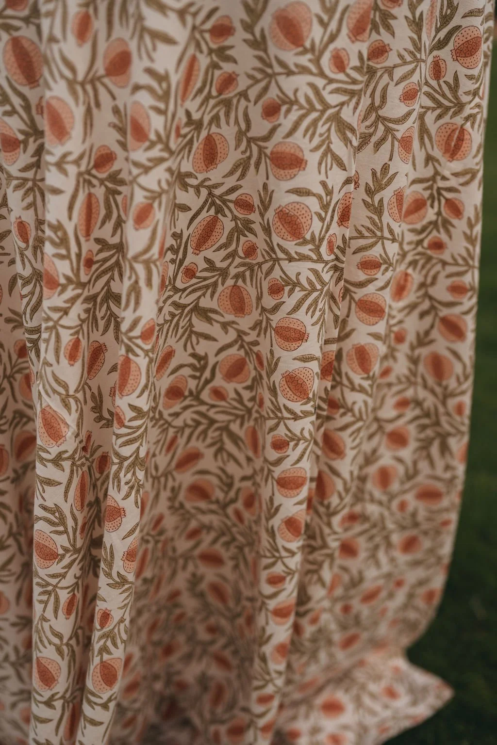 Close-up of a fabric with a floral pomegranate pattern featuring small orange flowers and brown leaves on a cream background.