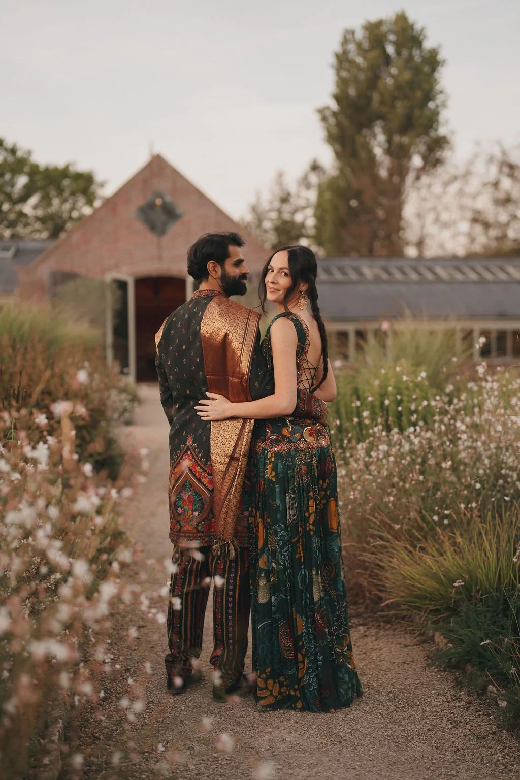 Couple in traditional Sikh wedding attire standing on a garden path with flowers, house, and trees in the background during sunset.