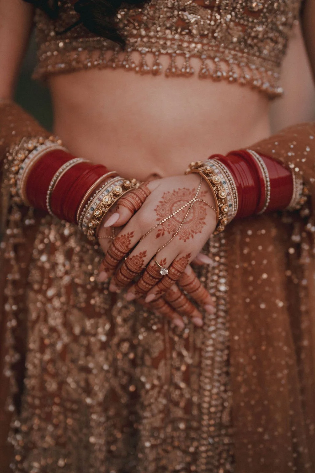 Close-up of traditional Indian bride's hands adorned with henna, jewelry, and red bangles, with intricate gold embroidery on the clothing.