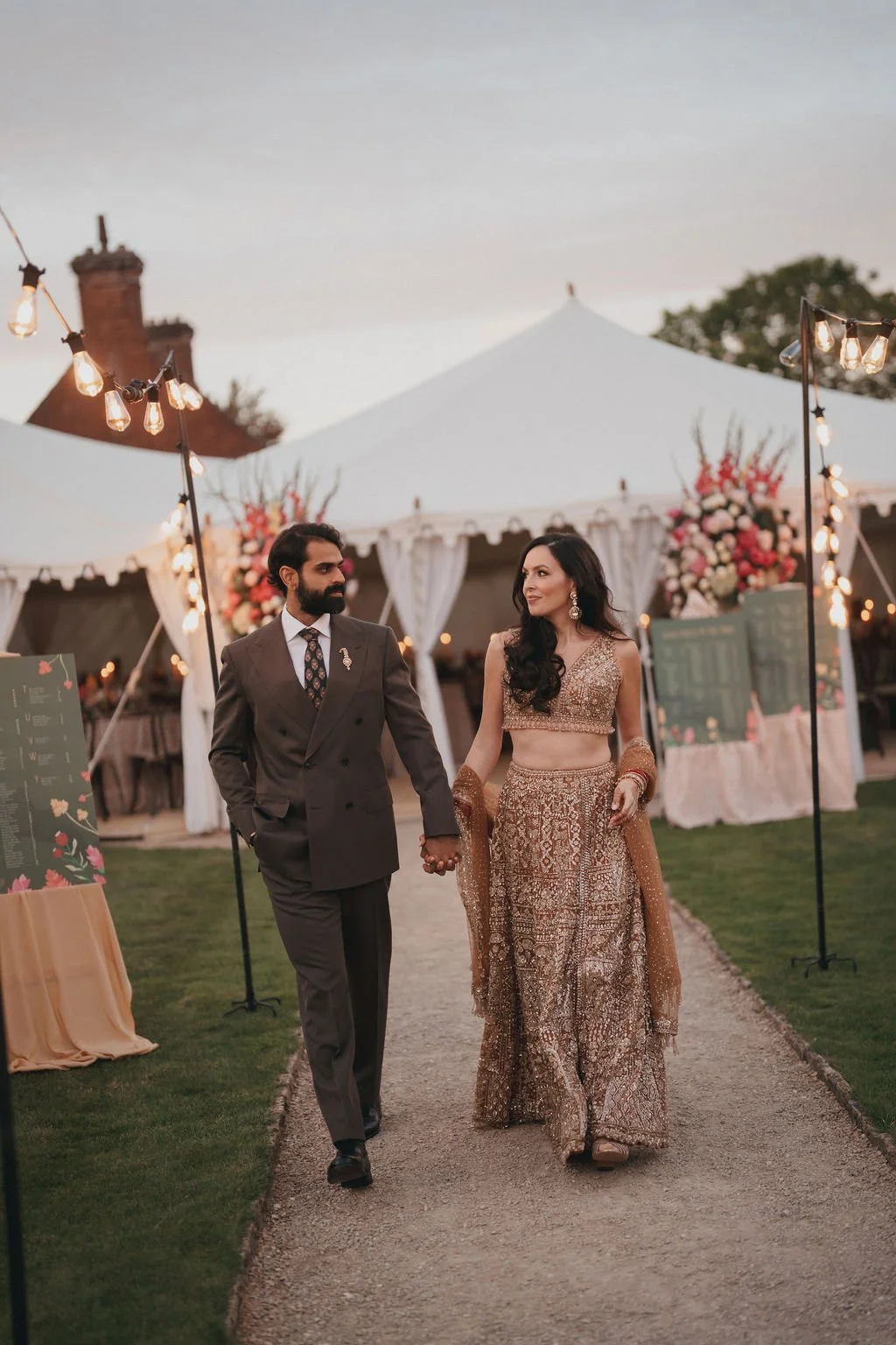 A couple walking hand in hand during an outdoor wedding reception at sunset. The woman is wearing a traditional Indian bridal outfit with intricate embroidery, and the man is dressed in a dark suit with a patterned tie. There are decorative lights, t
