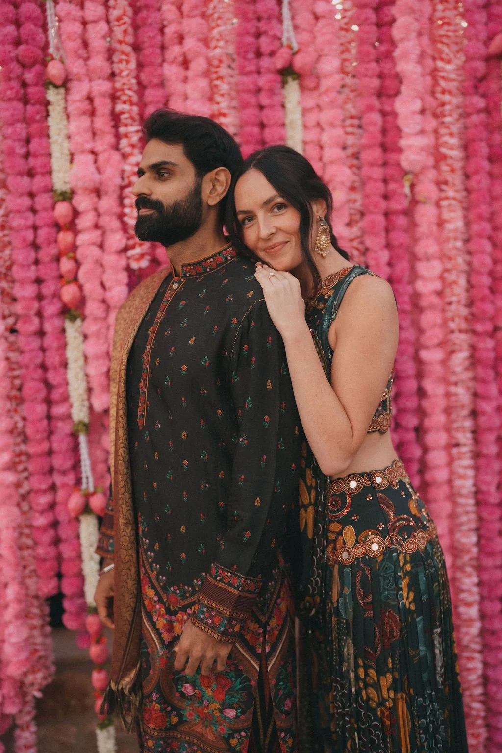 A bride and groom dressed in traditional Sikh wedding attire standing close together in front of a pink flower wall at Kin House.