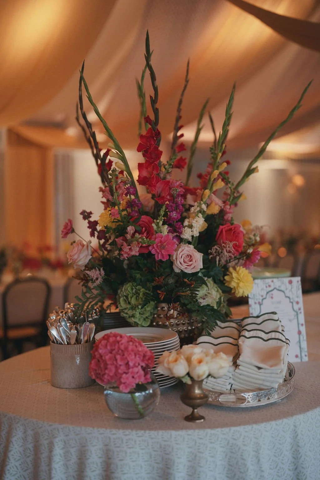 A large floral centerpiece with pink, yellow, and red flowers and green leaves on a decorated table. The table also has stacked plates, a small vase with pink flowers, a napkin holder with napkins, and a silver dish with white flowers. The setting ap