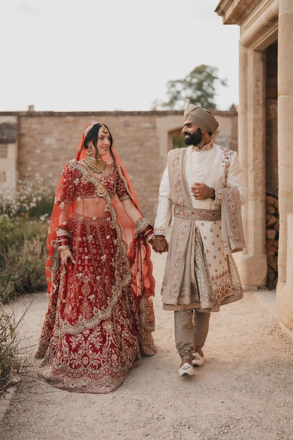 A bride and groom in traditional Indian wedding attire holding hands and smiling at each other outdoors.