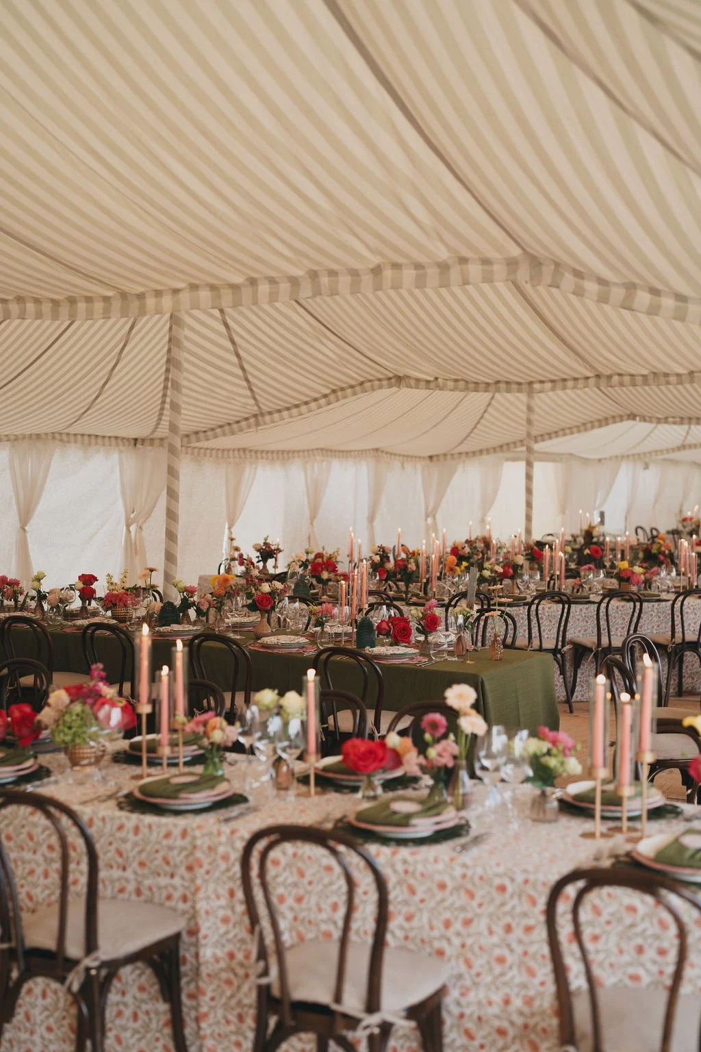Elegant wedding setup with long tables decorated with floral centerpieces and candles inside a large striped canopy marquee.