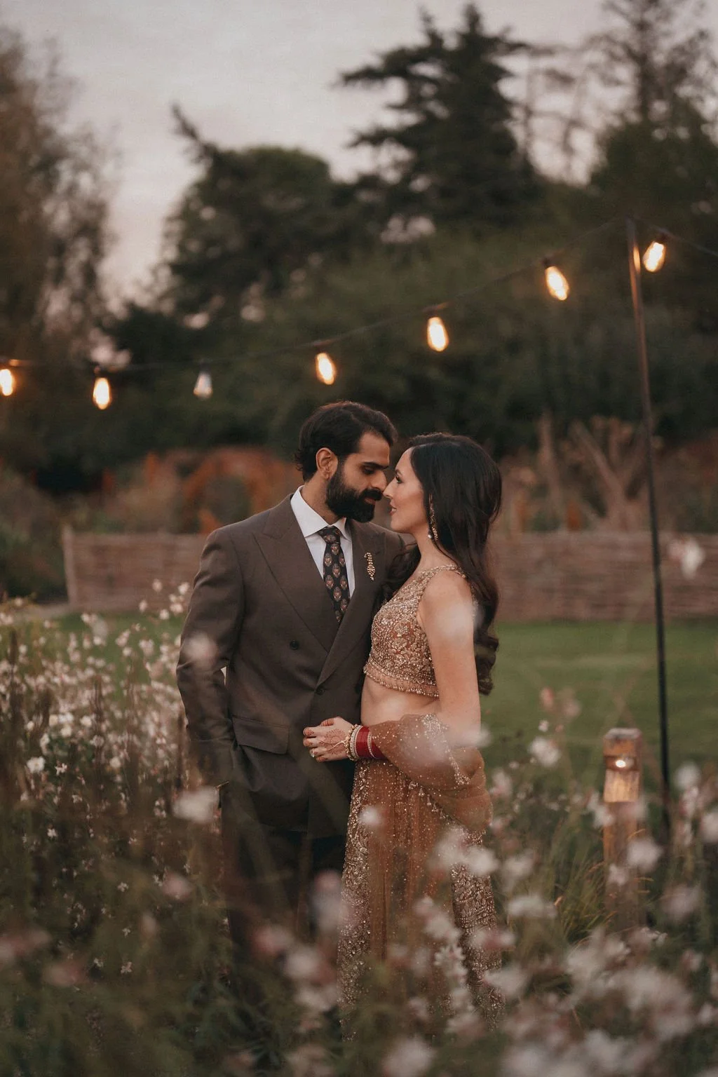 A couple dressed in traditional Indian wedding attire standing close together outdoors in a garden at dusk, surrounded by flowers and string lights.
