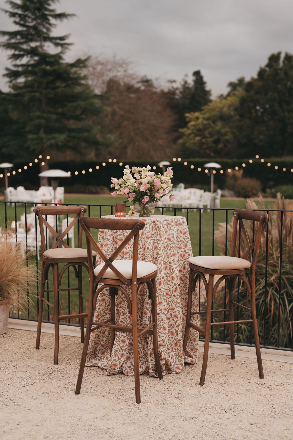 A table with a floral tablecloth and a flower arrangement in a glass vase, surrounded by three wooden chairs with cream cushions, set outdoors on a gravel surface with a black metal fence and string lights in the background.