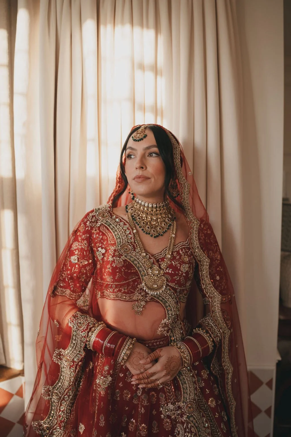 A woman dressed in a traditional red and gold Indian bridal outfit, adorned with heavy jewelry, henna on her hands, and a veil, standing in front of cream-colored curtains.
