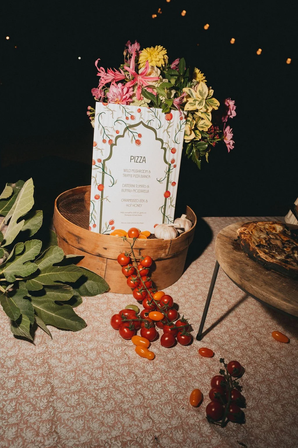 A table display with a basket of flowers, a tomato and garlic, a pizza on a round table, and a menu card listing pizza options. Decorative leaves are on the tablecloth.