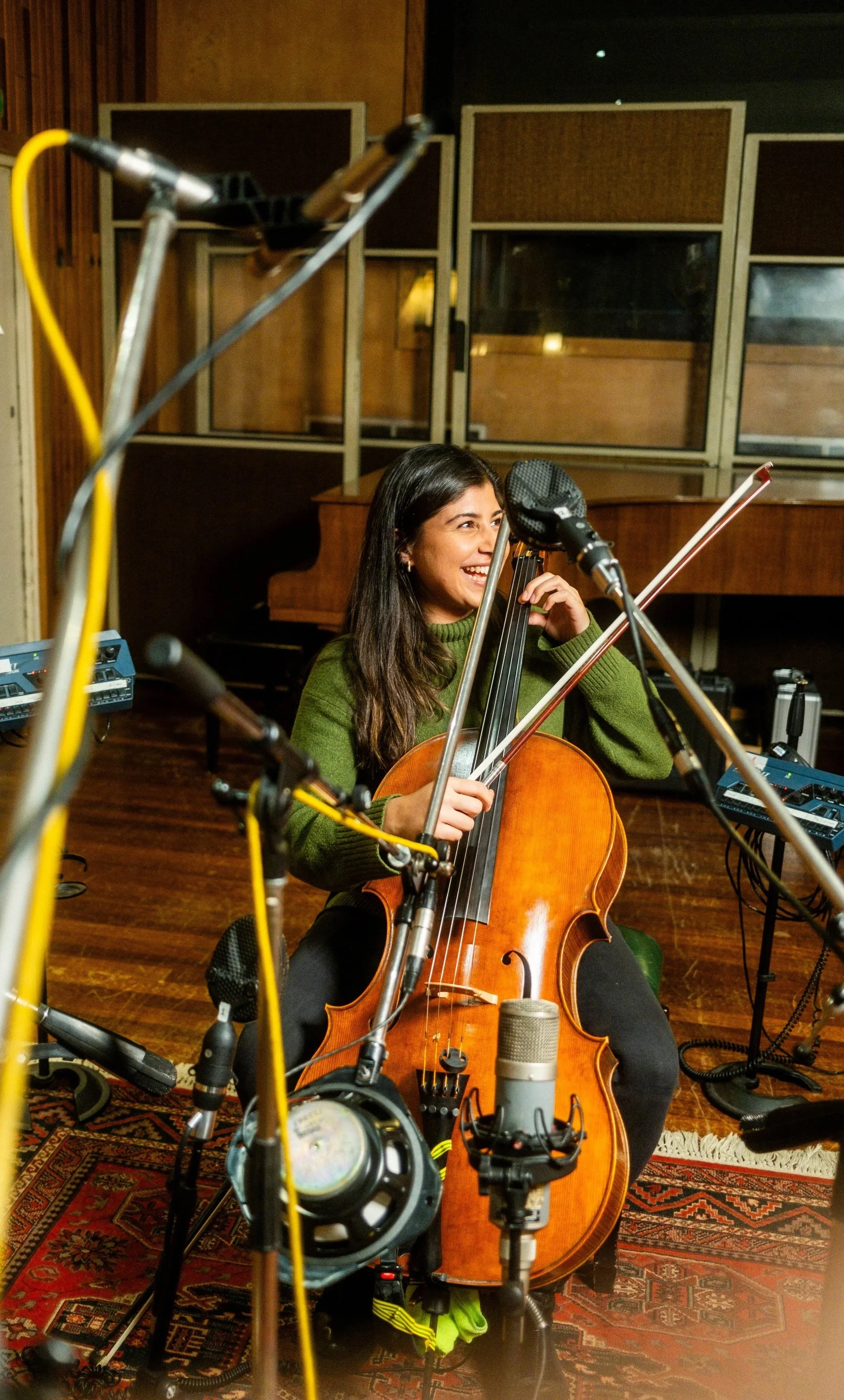 A woman playing the cello in a recording studio, surrounded by microphones and audio equipment, with a piano in the background.