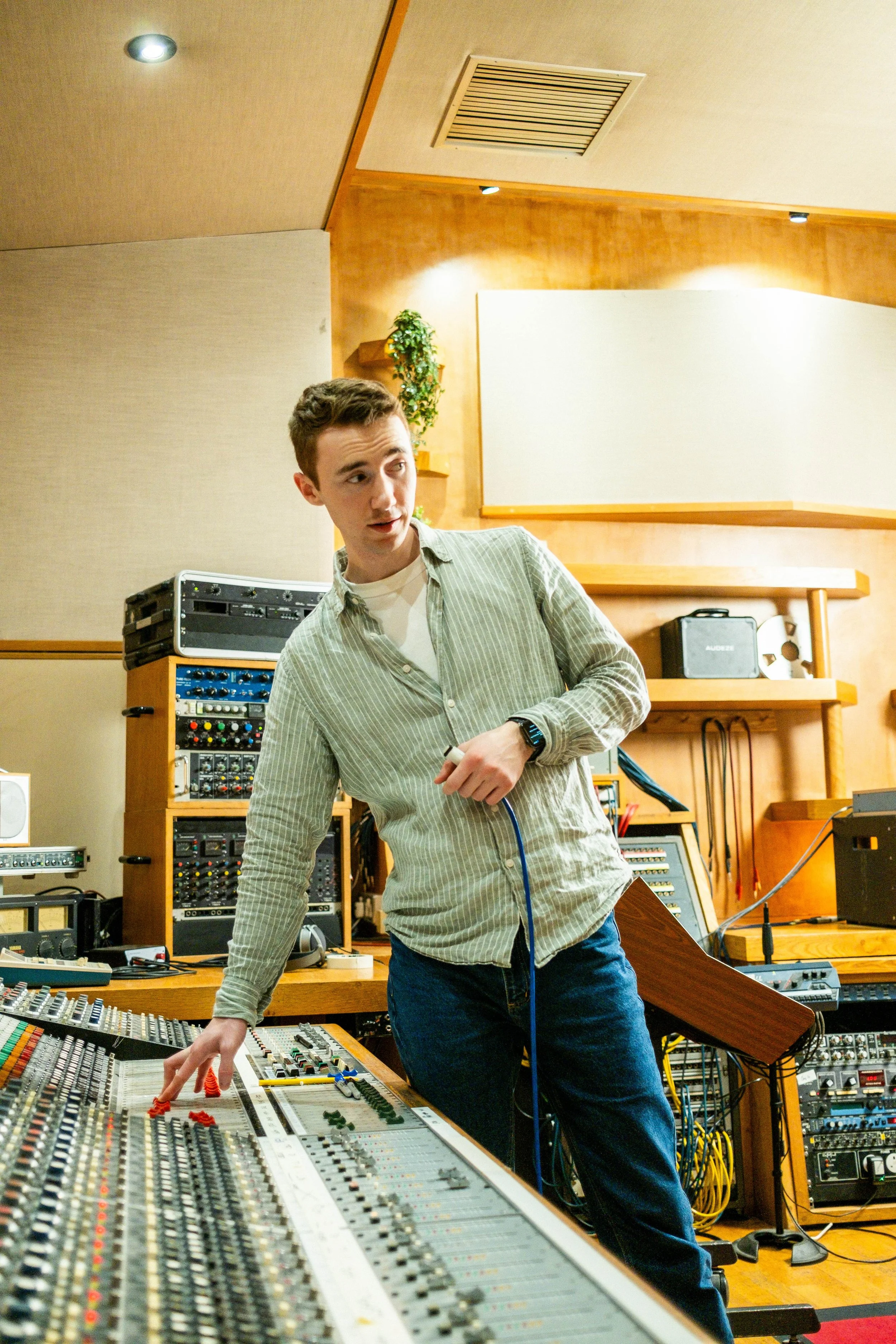 A young man wearing a striped shirt and jeans working at a large audio mixing console in a recording studio with various audio equipment and soundproofing panels.