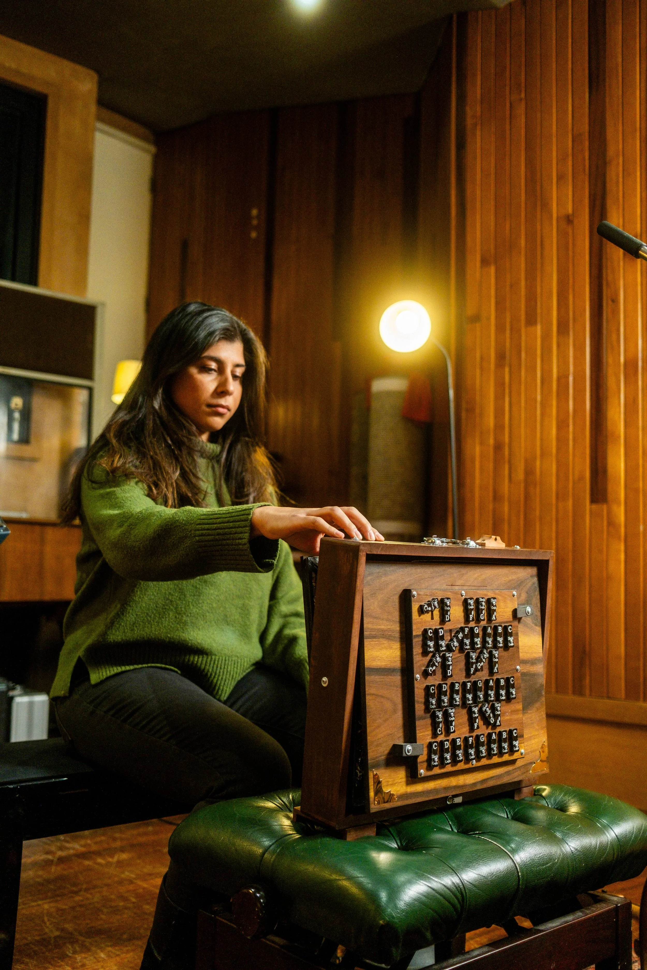 A woman with long dark hair wearing a green sweater playing a vintage wooden music box with black keys and white labels on a green leather tufted ottoman in a wood-paneled room with warm lighting.