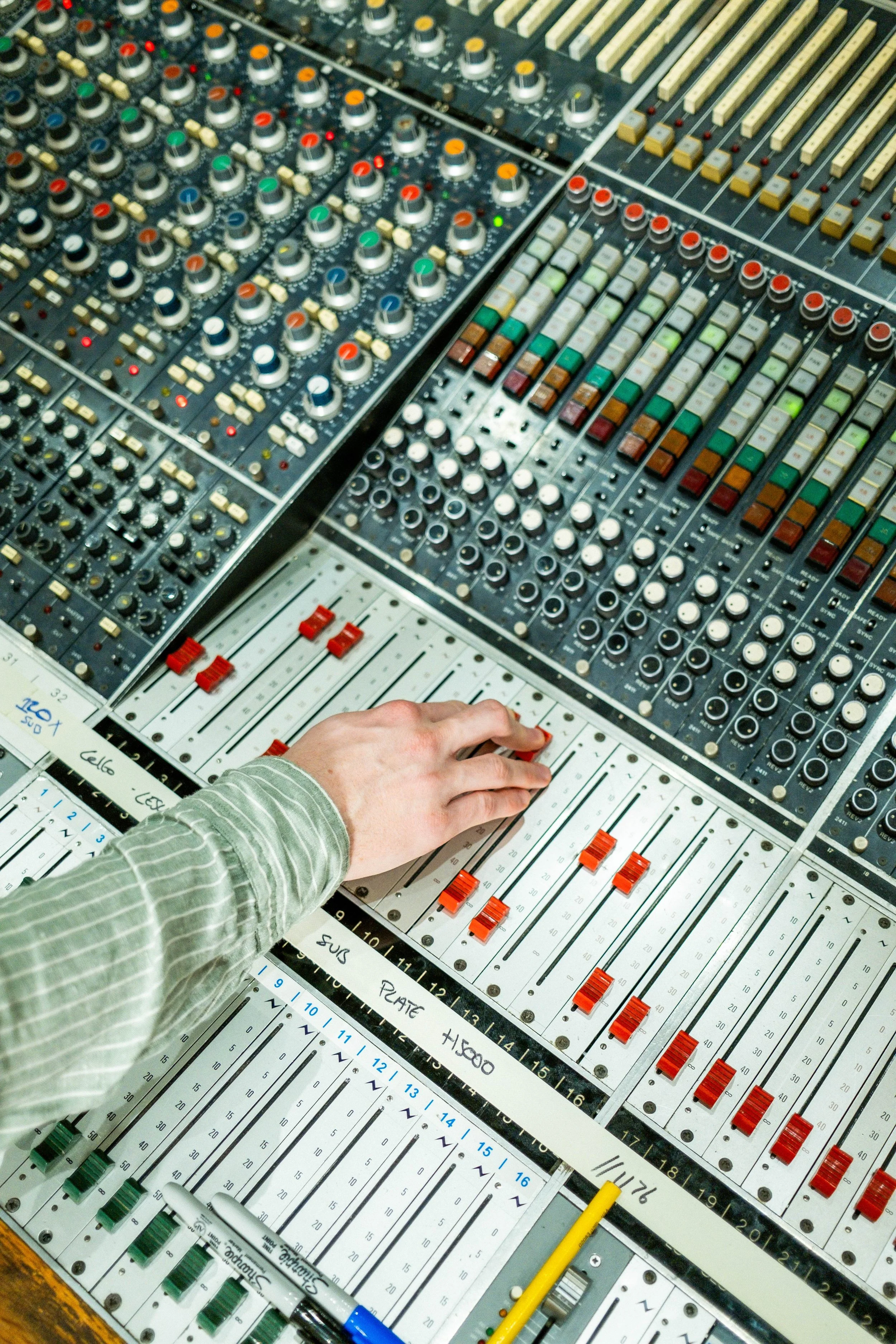 Close-up of an audio mixing console with numerous knobs, sliders, and colored buttons. A person's hand is adjusting one of the sliders, wearing a green and white striped long-sleeve shirt.