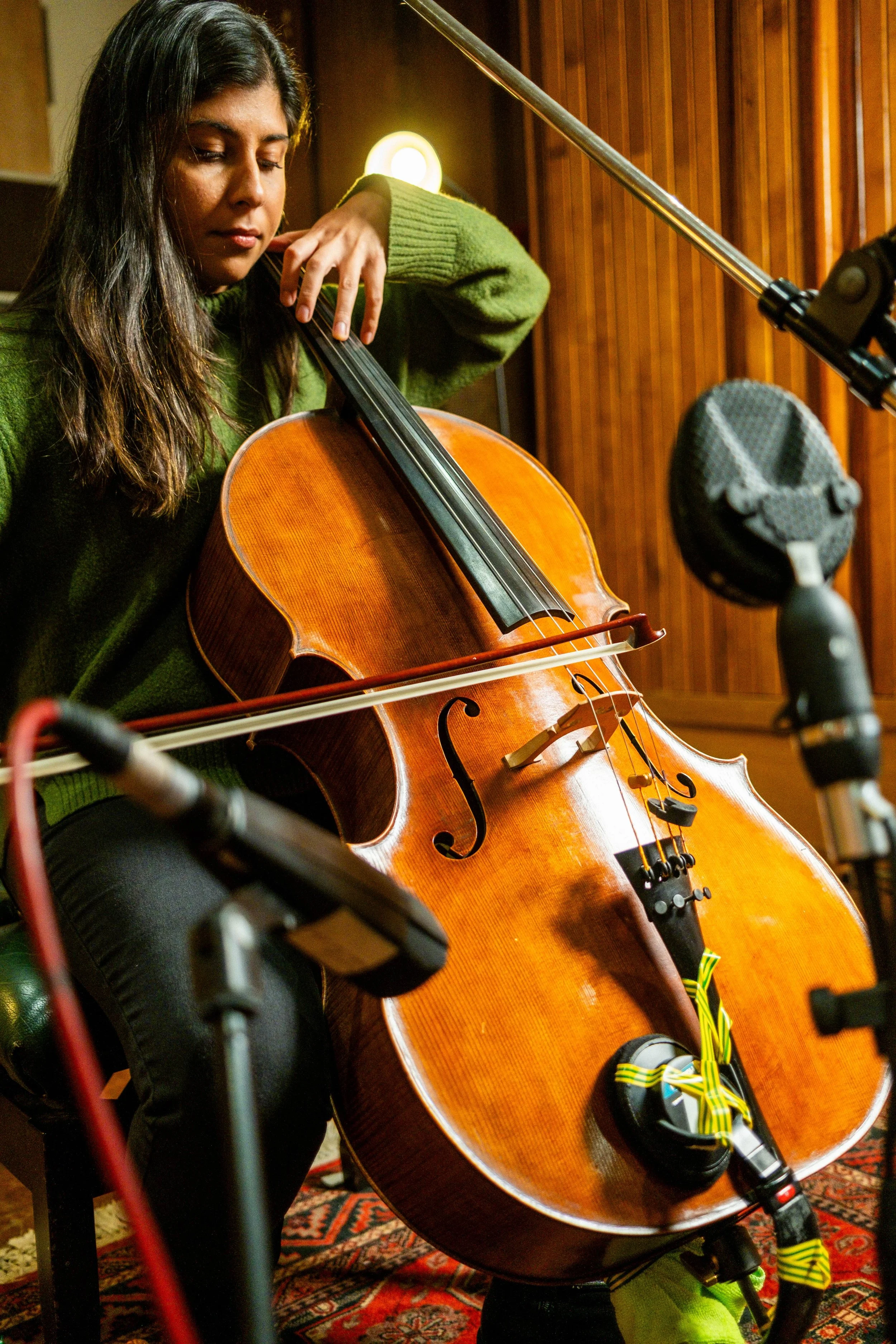 A woman with long dark hair playing a cello in a recording studio, with microphones and audio equipment around her.