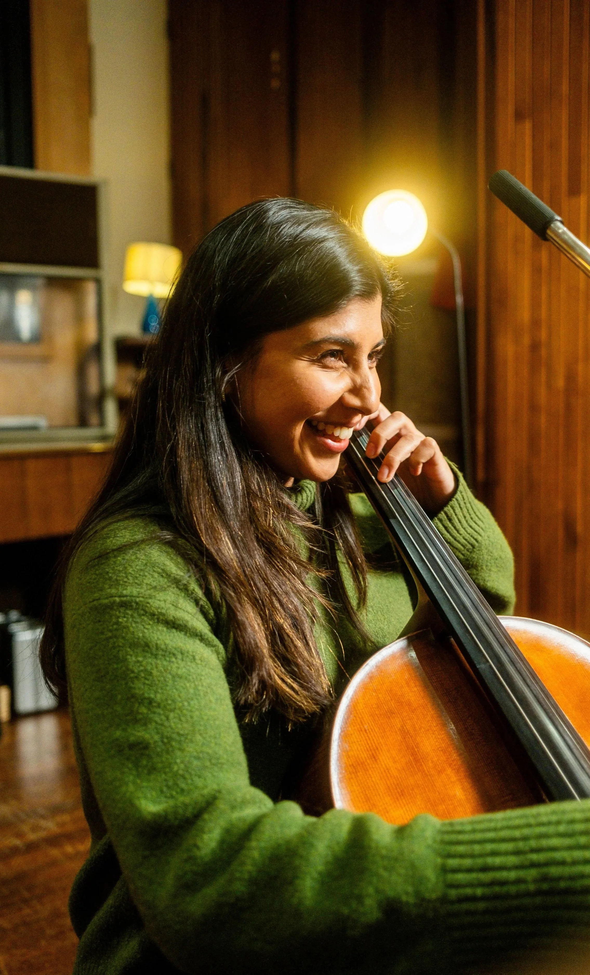A woman with long dark hair, smiling, playing the cello in a room with wooden walls and warm lighting.