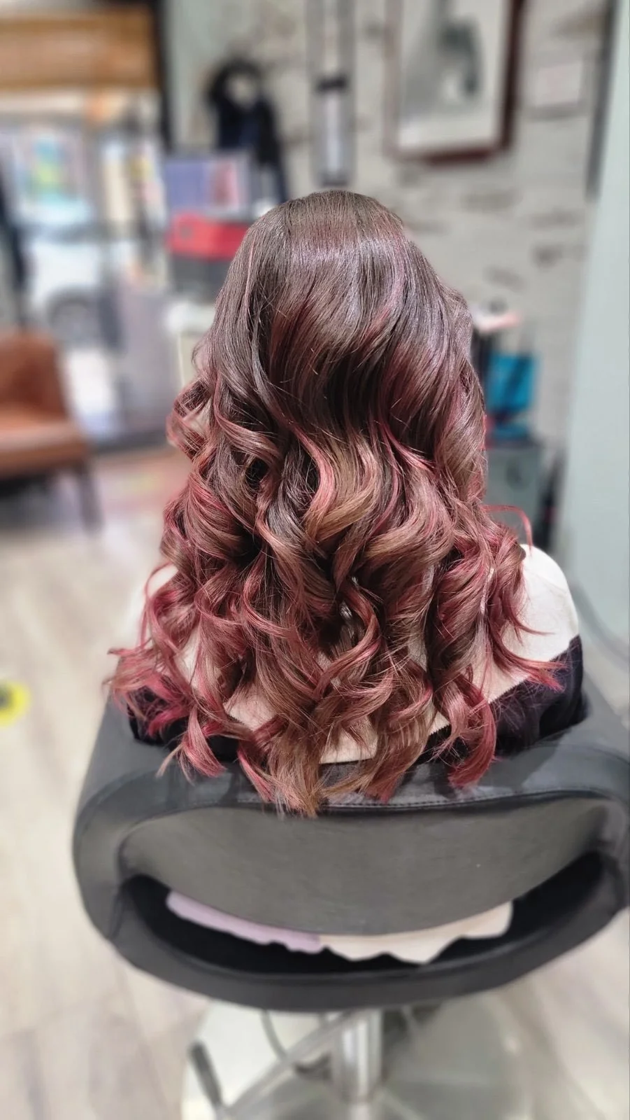 Back view of a salon client with long, voluminous wavy hair in a glossy burgundy-brown and pink ombré style.