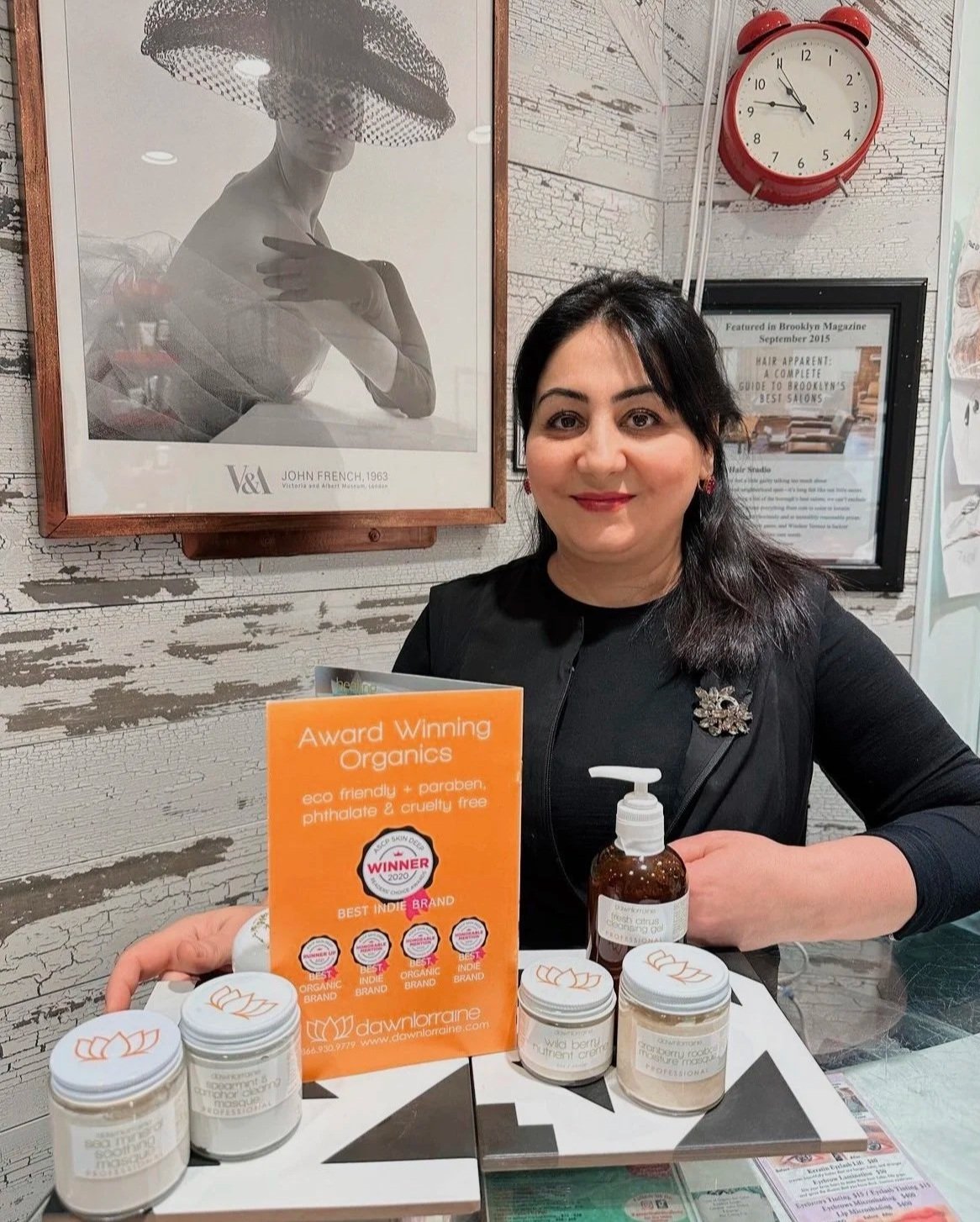 A woman with dark hair, wearing a black top and red earrings, sitting at a table with skincare products and a hand sanitizer. Behind her are framed pictures, including one of a woman wearing a hat, and a red clock showing 10:09. There is an orange sign promoting organic, cruelty-free skincare, and a clear wall with a textured, distressed finish.