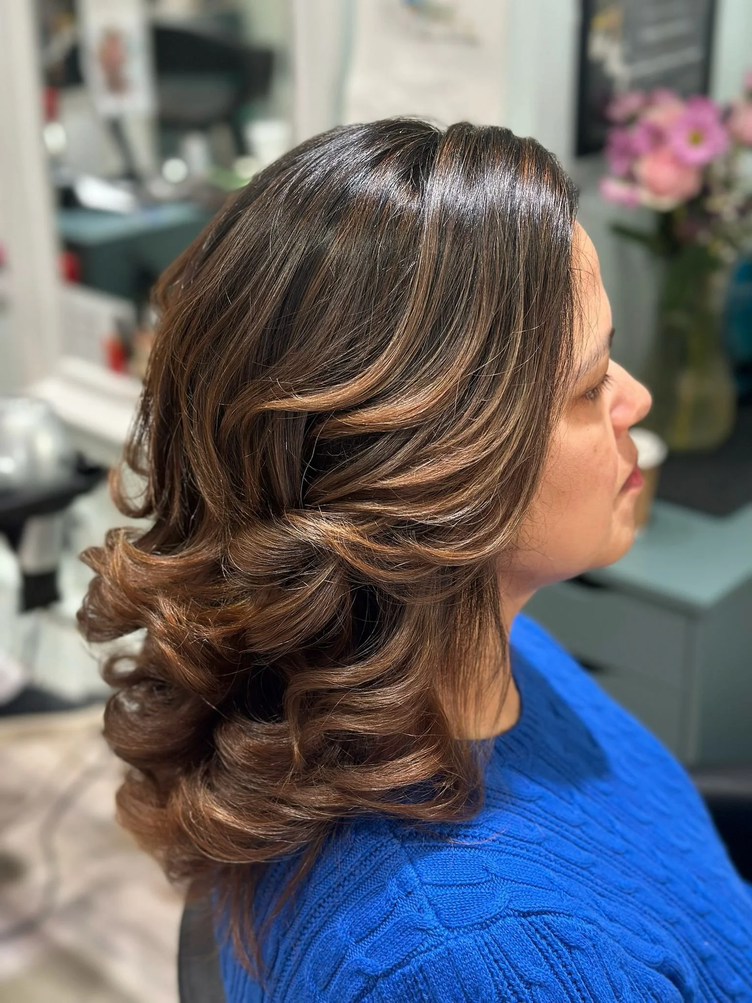 Woman in a salon with shoulder-length layered brunette hair styled in soft curls and caramel highlights