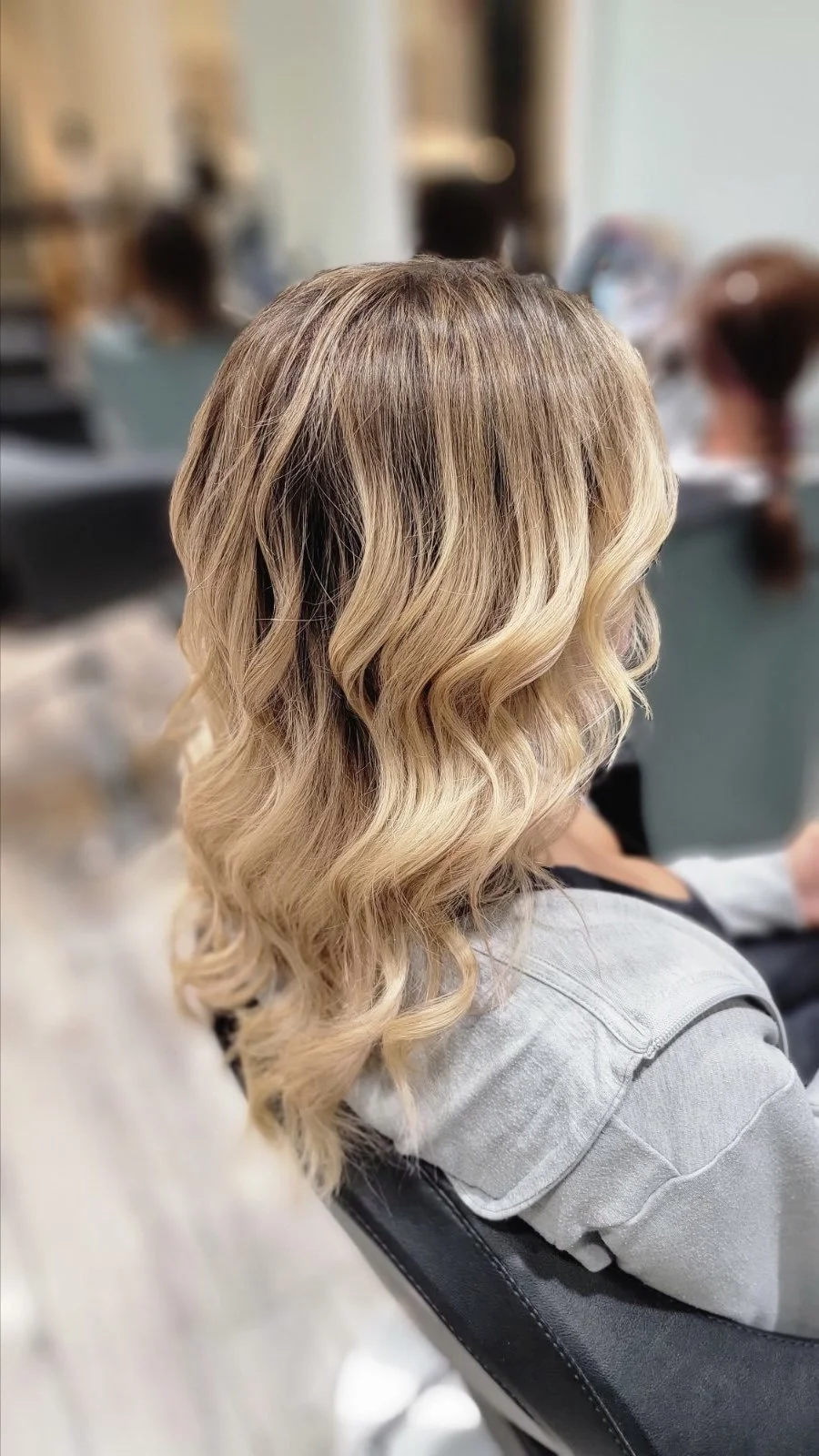Back view of a woman with shoulder-length blonde balayage hair styled in soft waves at a salon