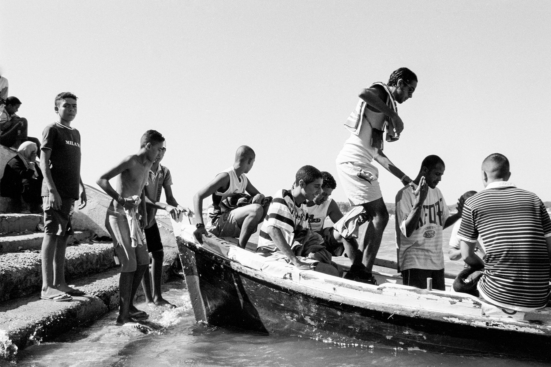 Group of young boys and men boarding a small boat at the water's edge on a sunny day.