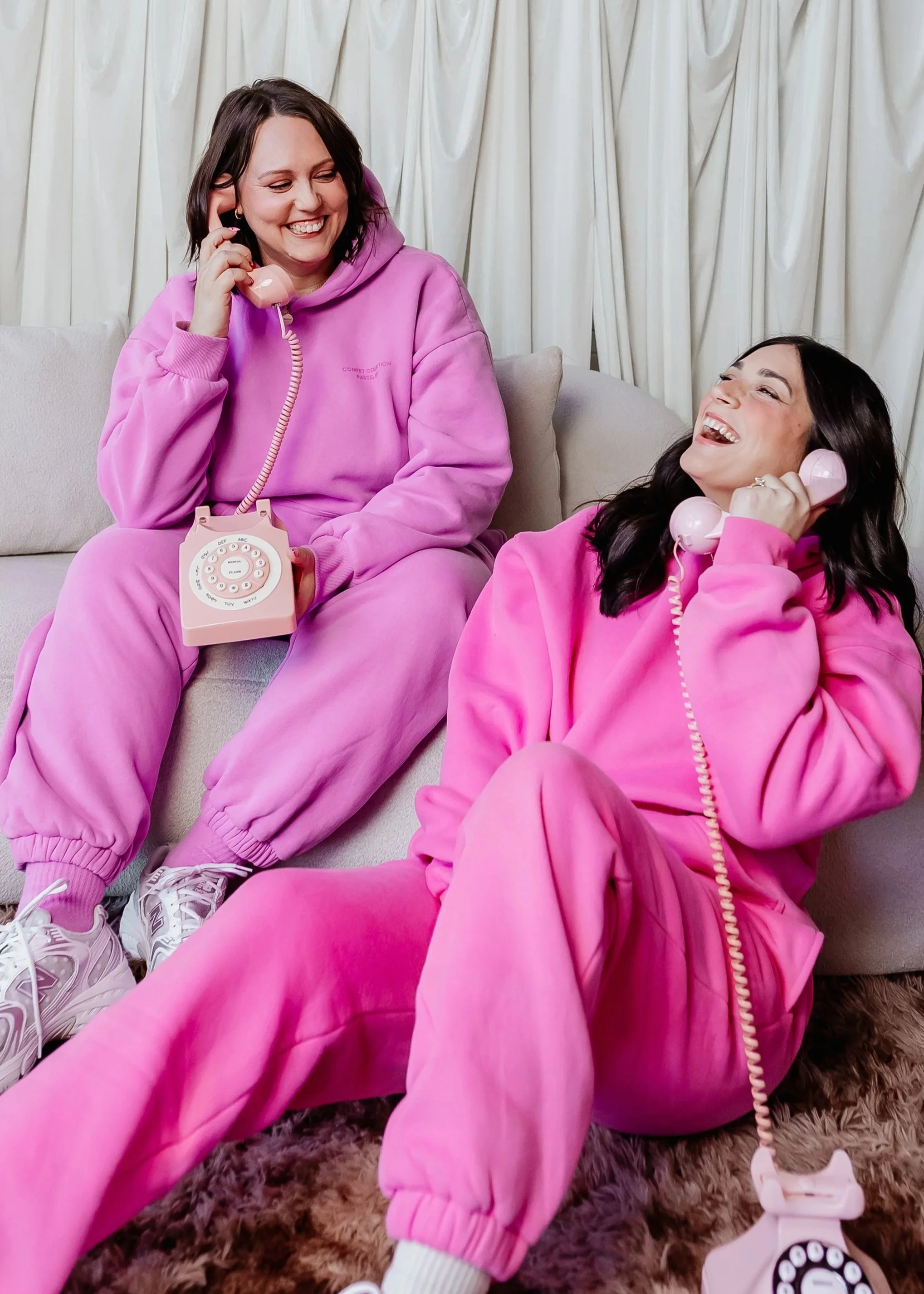 Two women in pink loungewear sitting on a sofa, talking on vintage pink rotary phones, laughing and smiling, with white curtains in the background.