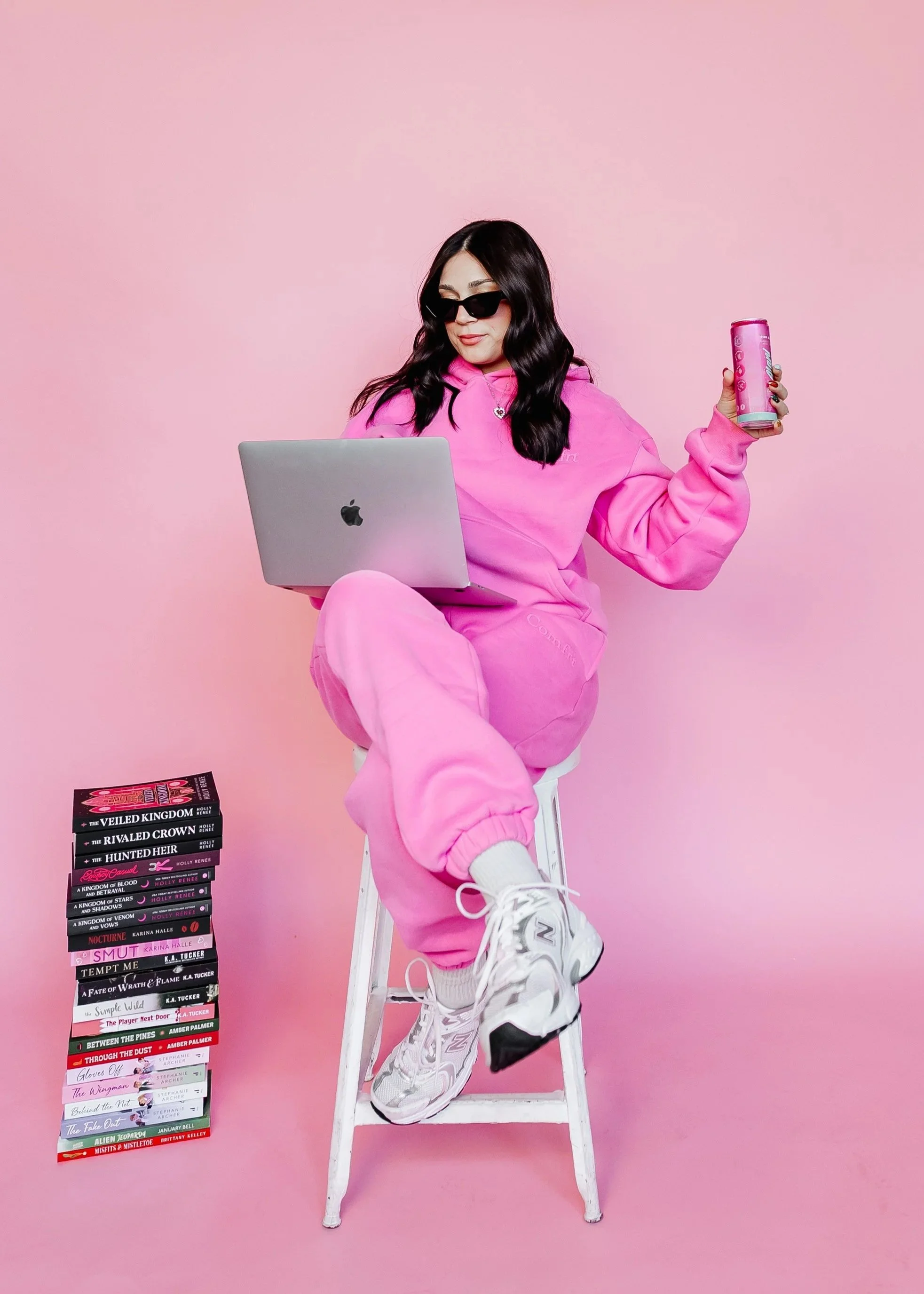 A woman wearing a pink hoodie and pink sweatpants is sitting on a white stool against a pink background. She is holding a can of soda in her right hand and a silver laptop in her lap. Next to her on the floor is a stack of books with titles related to royalty and fantasy, including "The Veiled Kingdom," "The Rivalent Crown," and "The Hunted Heir." She has long dark hair, sunglasses, and sneaker footwear.