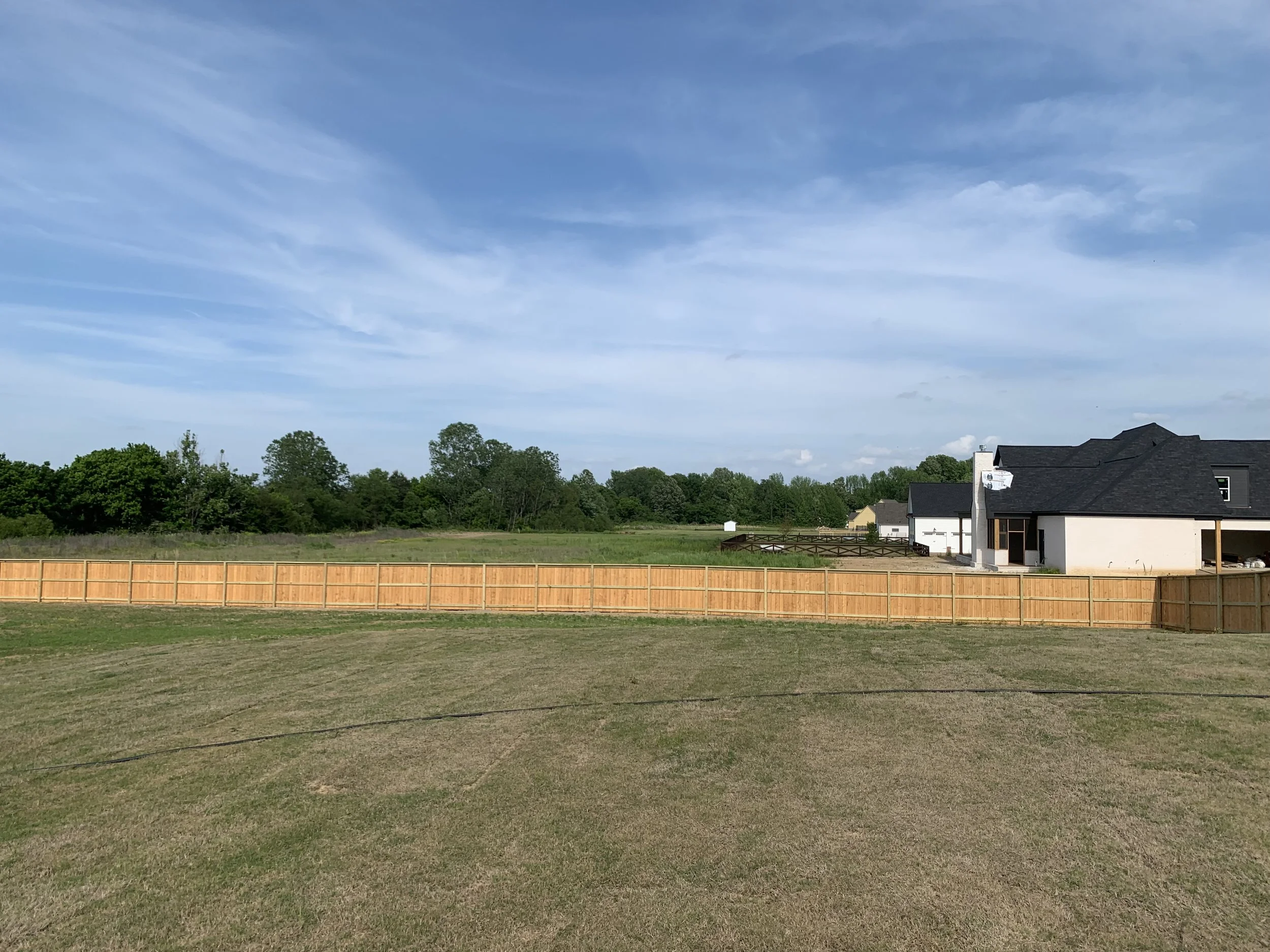 View of a backyard with a wooden fence, grass, trees in the distance, and a house with a black roof under a partly cloudy sky.