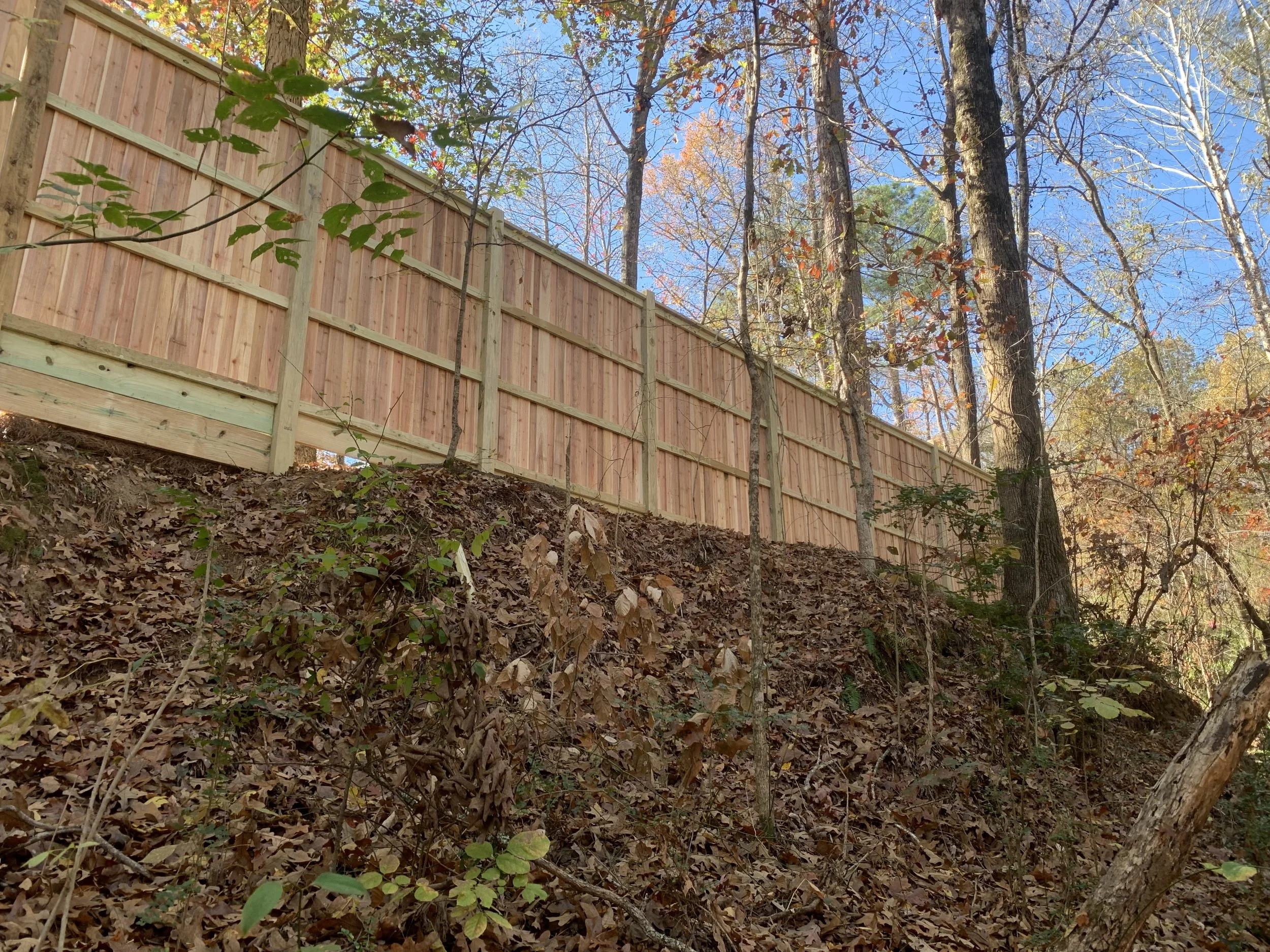 A wooden fence on a hill with autumn leaves and trees in the background, under a blue sky.