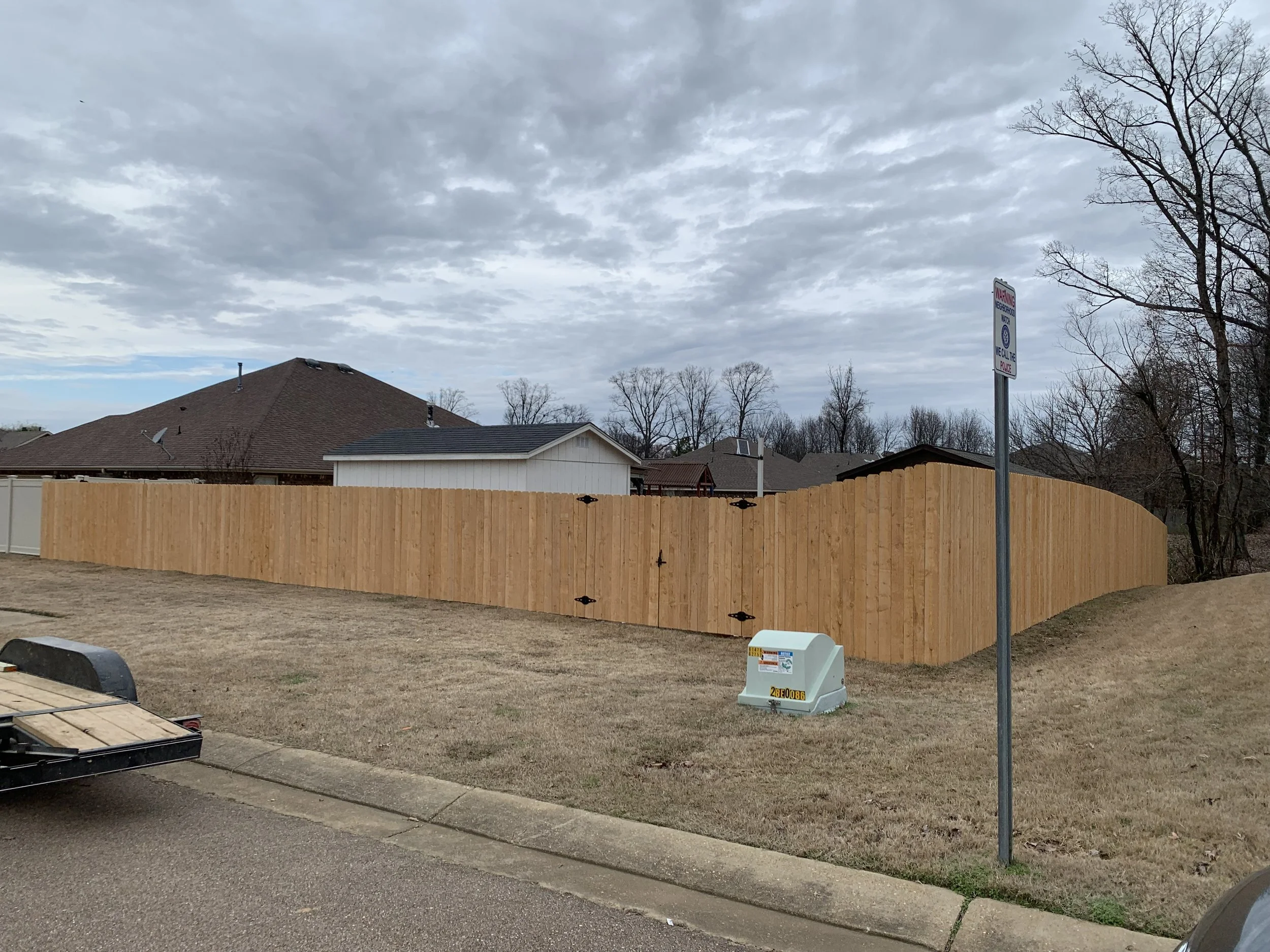 A backyard with a tall wooden fence, a white utility box, and a parking sign that says 'No Parking.' The sky is cloudy, and there are several houses and trees in the background.