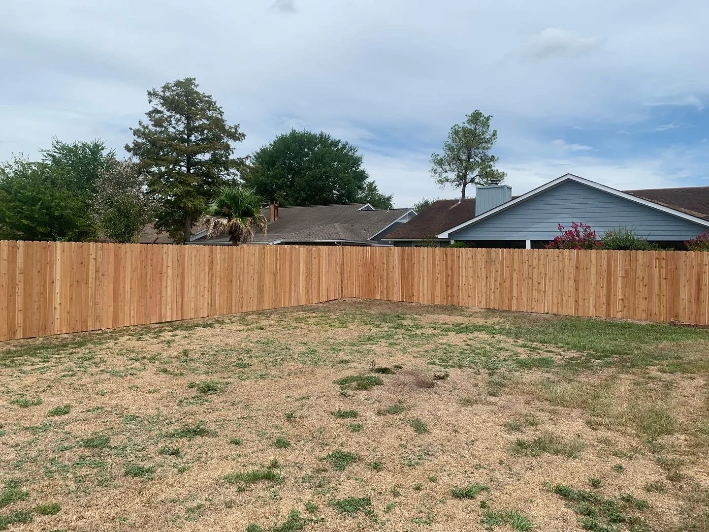 Backyard with a wooden privacy fence, patchy grass and dirt, neighboring houses with trees and shrubs, partly cloudy sky.