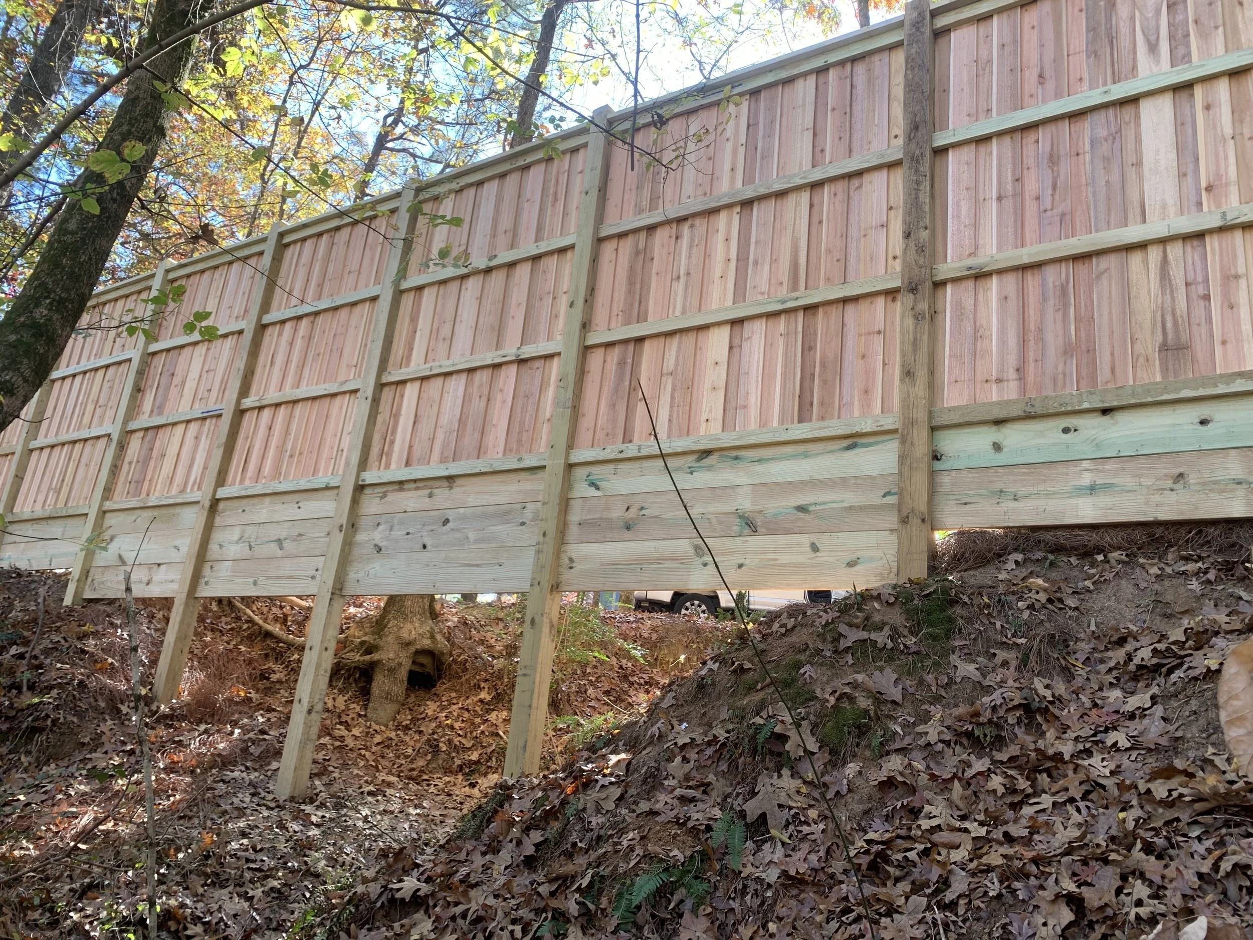 Wooden fence under construction on a hillside in a wooded area with trees and fallen leaves.