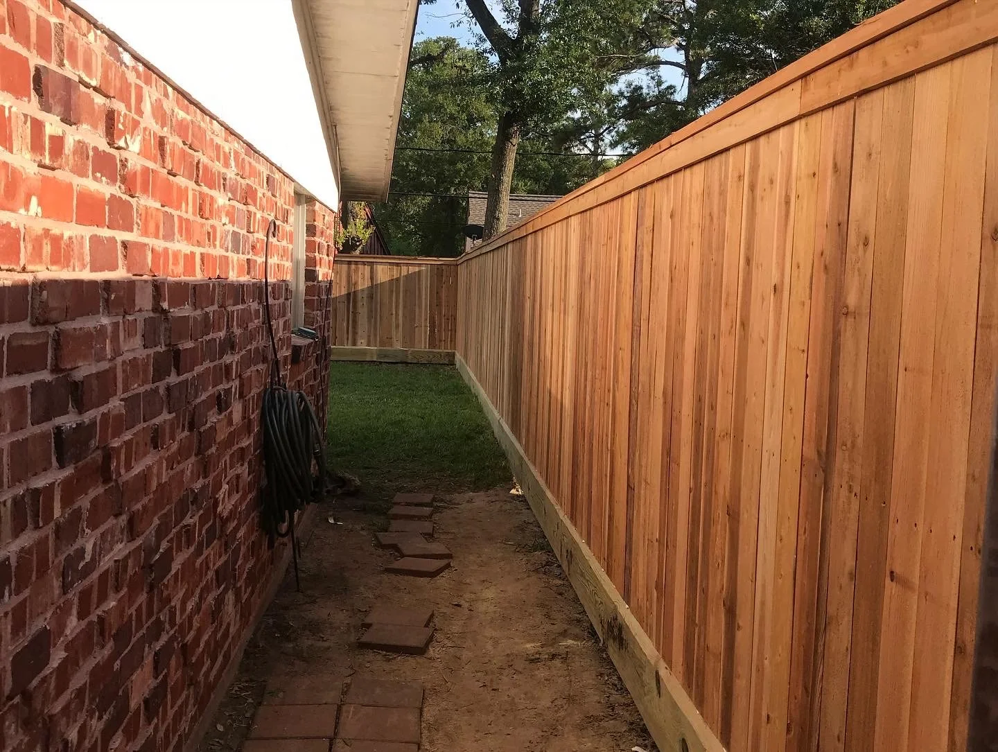 A narrow backyard space with a brick wall on the left and a tall, newly installed wooden fence on the right. There are paving stones on the ground leading towards the back. A garden hose is hanging on the brick wall, and there are trees and a house v