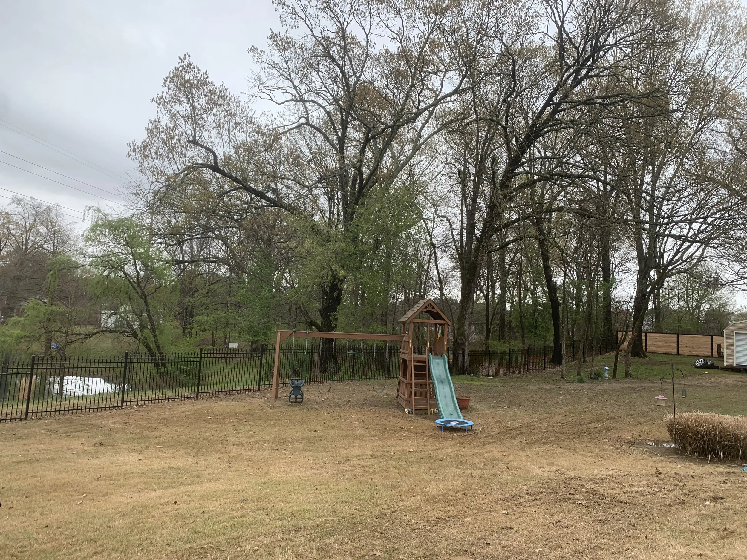 Backyard with a wooden playset, a slide, a trampoline, a chain-link fence, trees, and a cloudy sky.