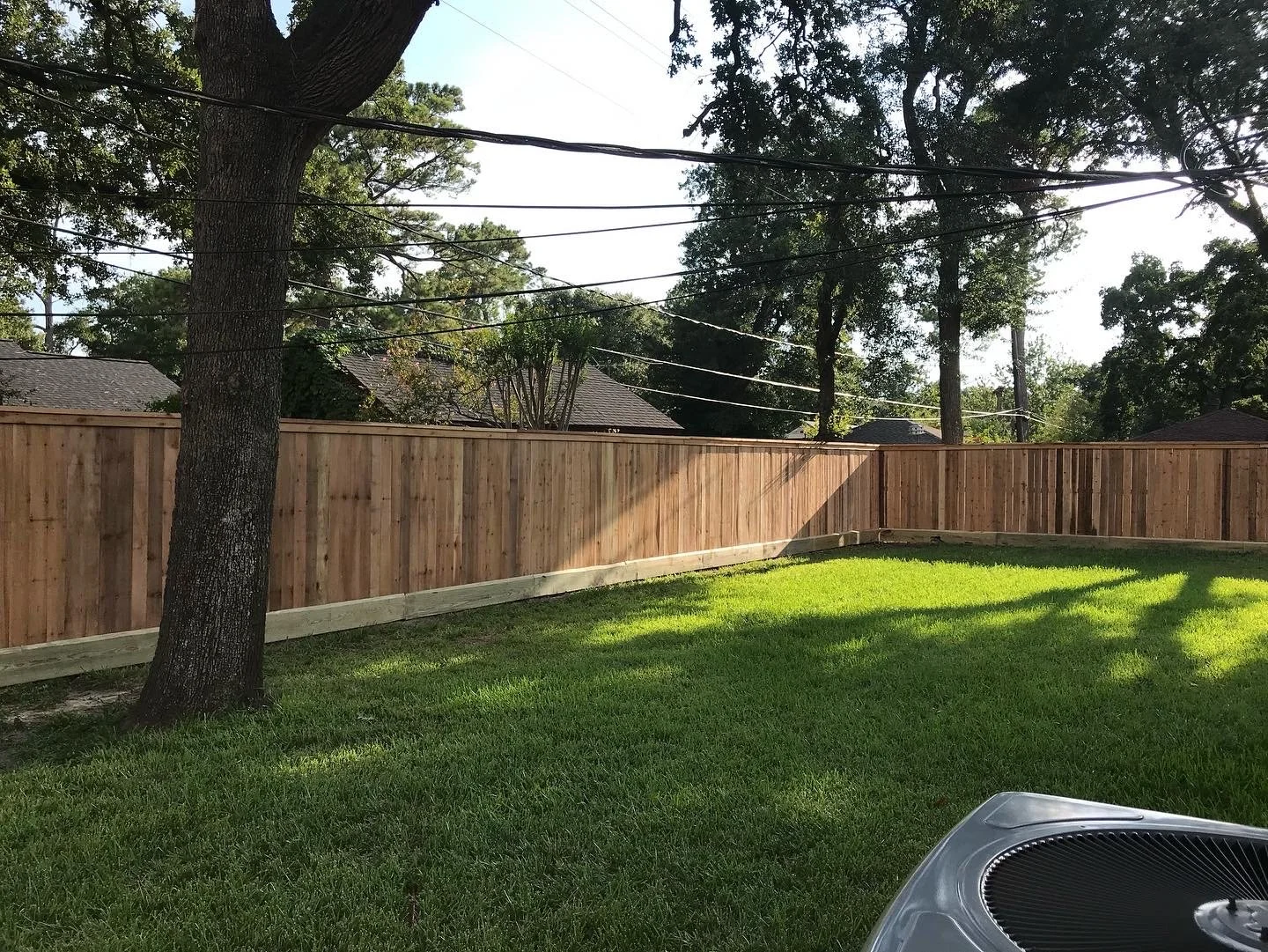 A backyard with a tall tree on the left, a wooden privacy fence, a grassy lawn, and a partial view of an air conditioning unit in the bottom right corner. Overhead power lines are visible in the background, along with a few rooftops and large trees.